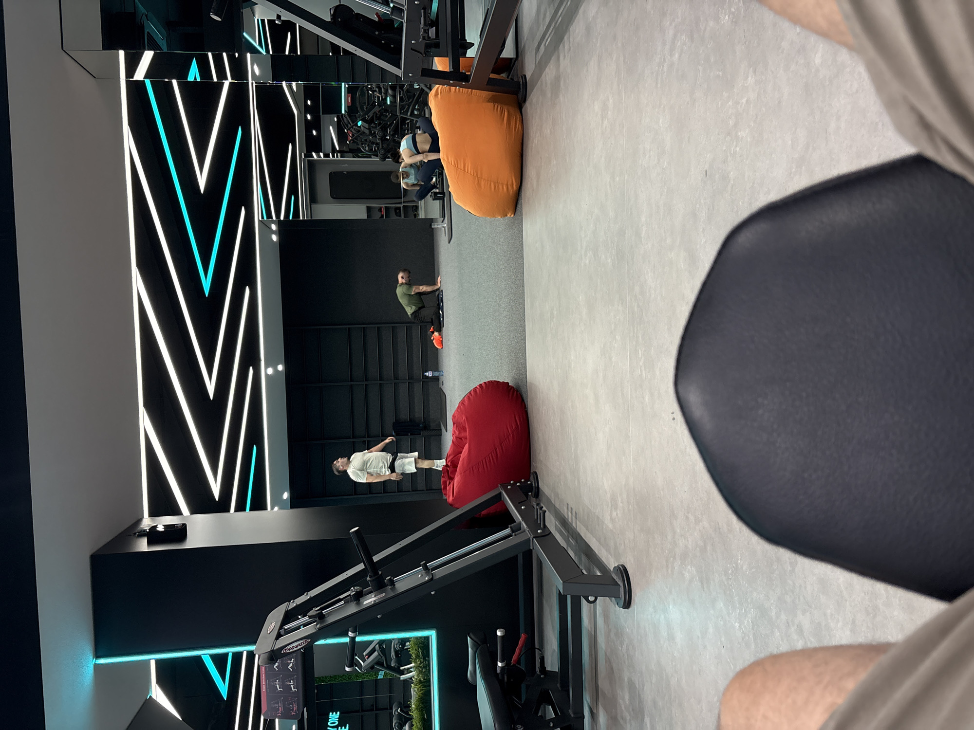 The image captures the interior of a modern gym, possibly located in Sofia, Bulgaria, indicated by the presence of gym equipment and a sleek design with neon lighting. The perspective is from a seated position, looking forward towards the gym floor.
In the mid-ground, several individuals are engaged in various activities. A man in a light-colored t-shirt and shorts stands near a large, red beanbag chair, appearing to be in the process of wiping sweat from his brow. Further back, another man in a green t-shirt is in a kneeling position on the floor, possibly performing a stretch or exercise. To his right, two women are visible, one in a light blue sports bra and leggings, and the other in a darker sports bra and leggings, both seated on the floor near an orange beanbag chair. Their reflections are visible in a large mirror.
The environment features a grey, textured floor and dark walls. The ceiling is adorned with a striking pattern of white and cyan neon lights, creating a dynamic and futuristic ambiance. A piece of weightlifting equipment, likely a leg press machine, is prominently positioned in the foreground on the left. Red and orange beanbag chairs are placed strategically on the gym floor, offering casual seating or rest areas. Behind the individuals, rows of black shelving units are visible, suggesting storage for gym accessories or equipment. The overall scene depicts a functional and aesthetically designed fitness space.