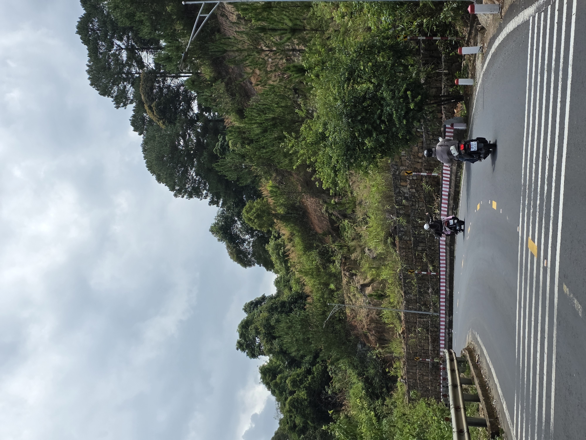 The image shows a winding road in Thôn 5, Vietnam. The road is paved and has white stripes painted on it, likely indicating a pedestrian crossing or a designated lane. A metal guardrail runs along the edge of the road. The road is bordered by a lush hillside covered in trees and vegetation. Two people are riding motorcycles on the road. The sky is overcast with grey clouds.