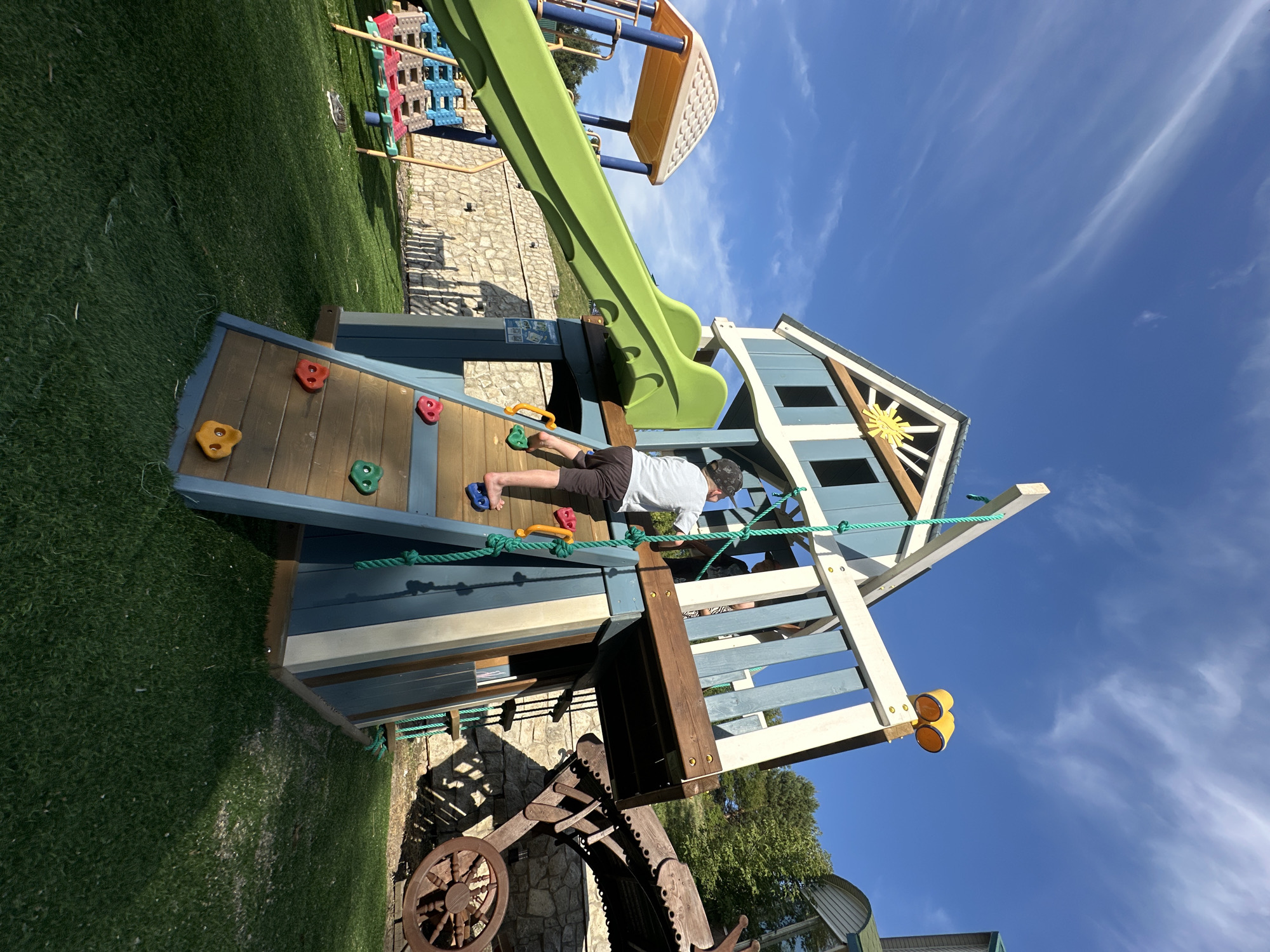 In an outdoor setting on a sunny day, a young boy wearing a grey t-shirt and dark shorts is climbing a blue and white wooden playset. The playset has a green slide, a climbing wall with colorful handholds, and a rope. Behind the playset, there is a stone wall, green artificial turf, trees, and a clear blue sky with wispy white clouds. In the background, a wooden wagon wheel and part of a wooden structure are visible. The location is identified as Danilovka, Altai Krai, Russia.