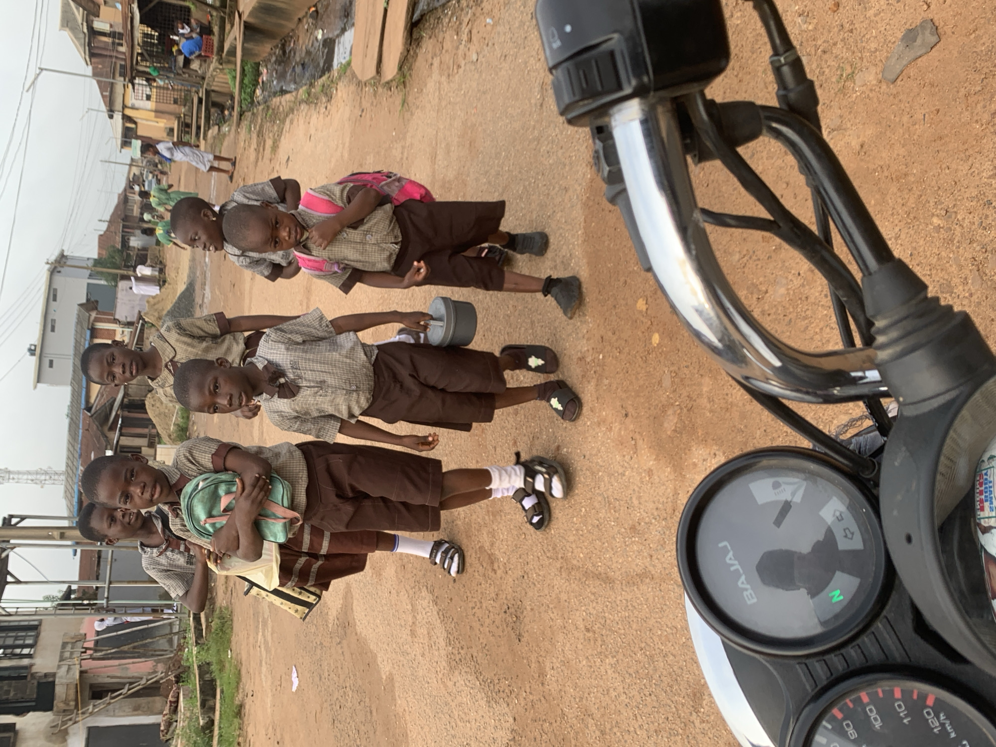 In Shagamu, Nigeria, a group of five school-aged children, dressed in uniform shirts and shorts, walk along a dirt road. The children are carrying backpacks and bags, and one child holds a small metal container. The road is lined with buildings, some appearing to be residential or shops. In the foreground, a motorcycle's handlebars and dashboard are visible, suggesting the photograph was taken from the perspective of a rider. The environment appears to be a typical street scene in a developing urban area, with uneven terrain and visible drainage channels.