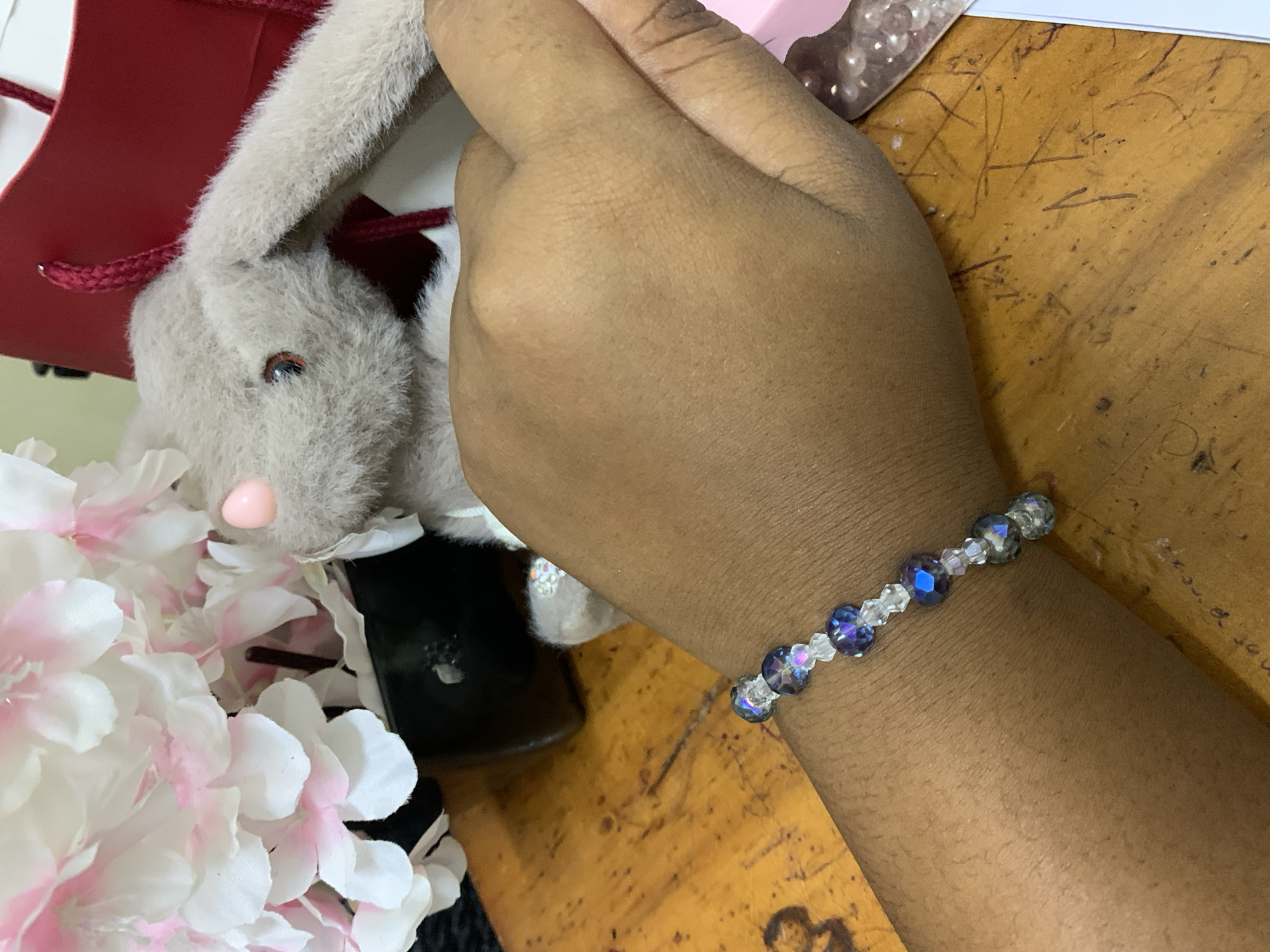 A right forearm with a bracelet is positioned diagonally on a wooden surface. The bracelet alternates between small, translucent beads and larger, dark blue beads. A stuffed grey rabbit with a pink nose and black eyes is in the background. To the left of the rabbit are pink and white artificial flowers. The rabbit appears to be next to a red and white bag. A pink container and clear beads are visible in the upper right. The wooden surface has scratches. It is not possible to identify the location in Dar es-Salaam, Tanzania, from the image.