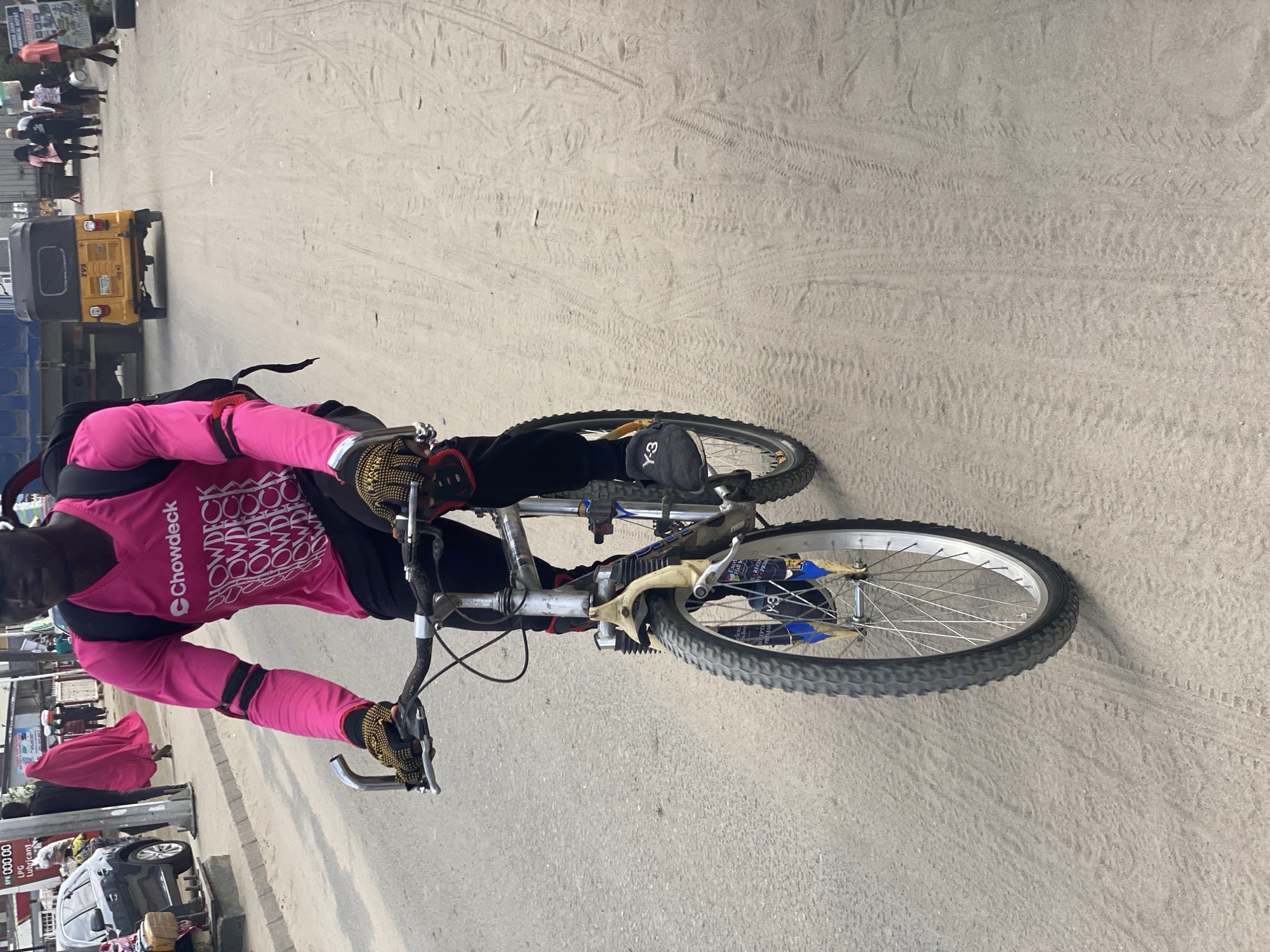 The image captures an outdoor street scene in Ado, Nigeria, likely during the daytime under bright, clear weather conditions. The ground is a mix of sandy patches with visible tire tracks and paved asphalt/concrete.
The central subject is a person riding a bicycle. They wear a bright pink long-sleeved shirt with the "Chowdeck" logo repeated prominently across the chest, black pants, and a black backpack. Gold-patterned cycling gloves are visible on their hands gripping the handlebars. A black fabric item bearing the "Y-3" logo is seen near their right knee, possibly a knee pad or a component of their gear. The bicycle is a silver/grey mountain bike style with black tires, silver spokes, and blue accents on the front fork, which also shows partial text like "Excel" and "REO PR" on stickers.
In the background, a yellow auto-rickshaw with "TVS" visible on its rear is parked or moving on the street. Further back on the left, several pedestrians are visible, some walking, others standing, contributing to a busy street atmosphere. One person in the distance is wearing a flowing red garment. A dark-colored car is partially visible on the far left.
The overall activity suggests a delivery rider on duty for "Chowdeck," navigating a bustling urban environment. Other text elements include "DPK 000 00" and "LPG Lubricant" on signs in the lower left background.