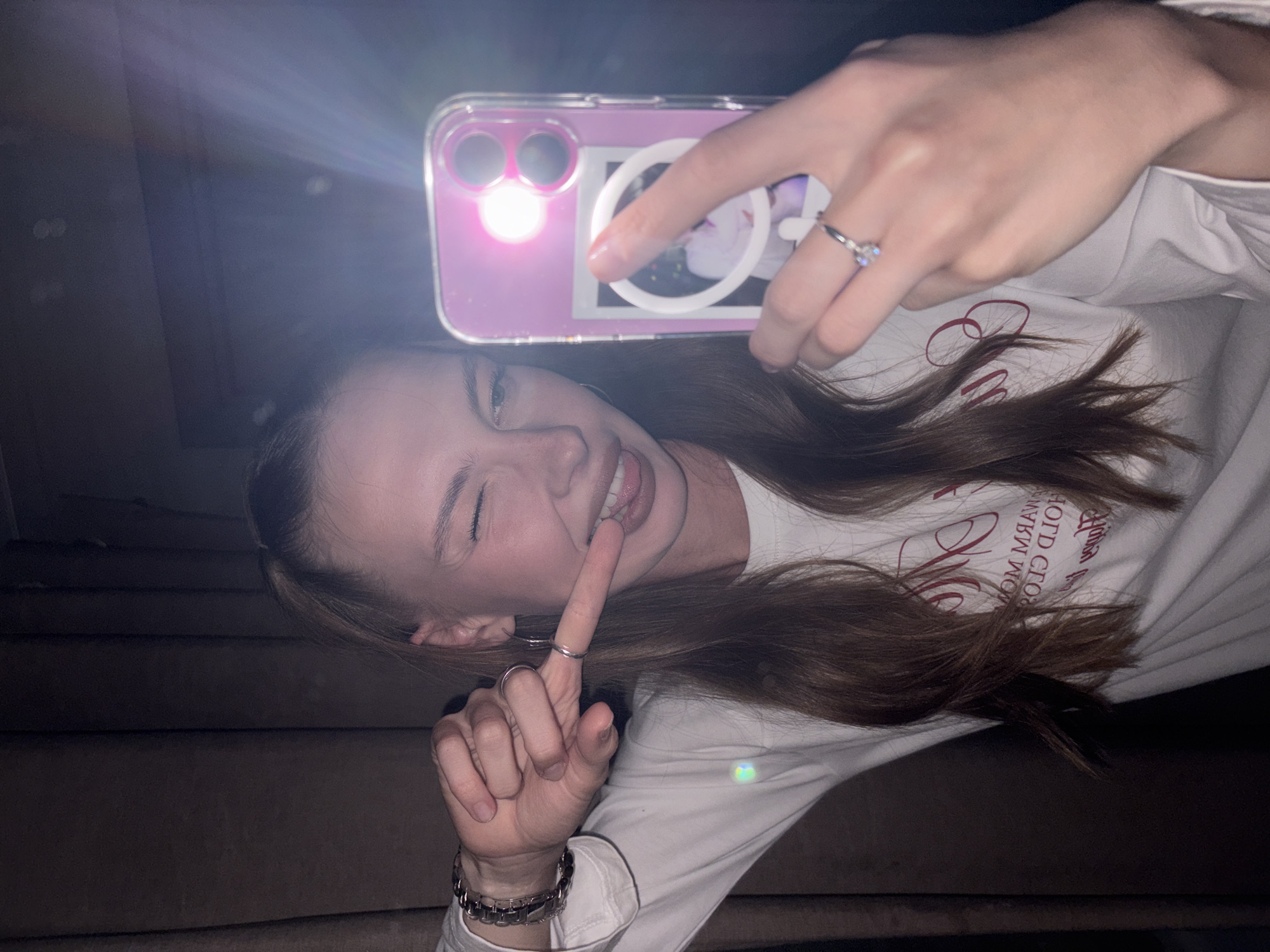 A young woman with long brown hair tied back in a ponytail is taking a selfie in a dimly lit room. She is wearing a long-sleeved white shirt with red lettering. Her right hand is holding a pink phone, which is in a clear case with a small picture inside. She is winking and sticking out her tongue, with her left index finger touching her tongue. A silver bracelet is visible on her left wrist, and she is wearing a ring with a diamond on her right hand. The background appears to be a dark curtain. The image does not provide any specific details that would indicate a location in the city of Perm, Russia.