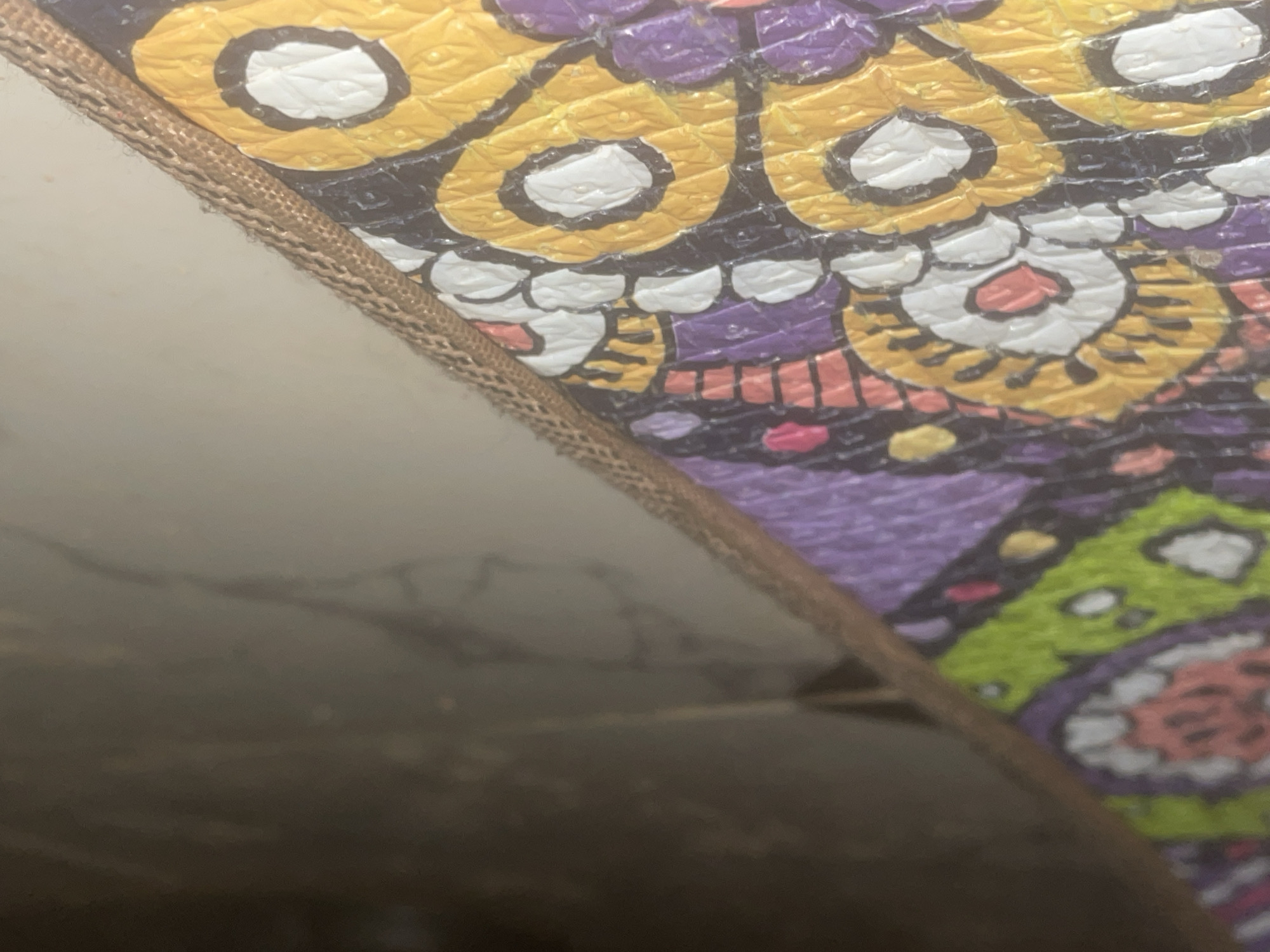 A close-up, eye-level, indoor shot shows a portion of a colorful, patterned rug with a woven brown trim along its right edge. The rug features a repeating mandala-like design with concentric circles in yellow, purple, green, and pink, outlined in black and white. The surface of the rug appears textured, with a slightly glossy finish. The background consists of a blurred, neutral-toned floor, possibly marble, and a dark, out-of-focus area above. There is no indication of the city of Howayi, Nigeria, or any specific location. The image focuses solely on the detail and texture of the rug and its immediate surroundings.
