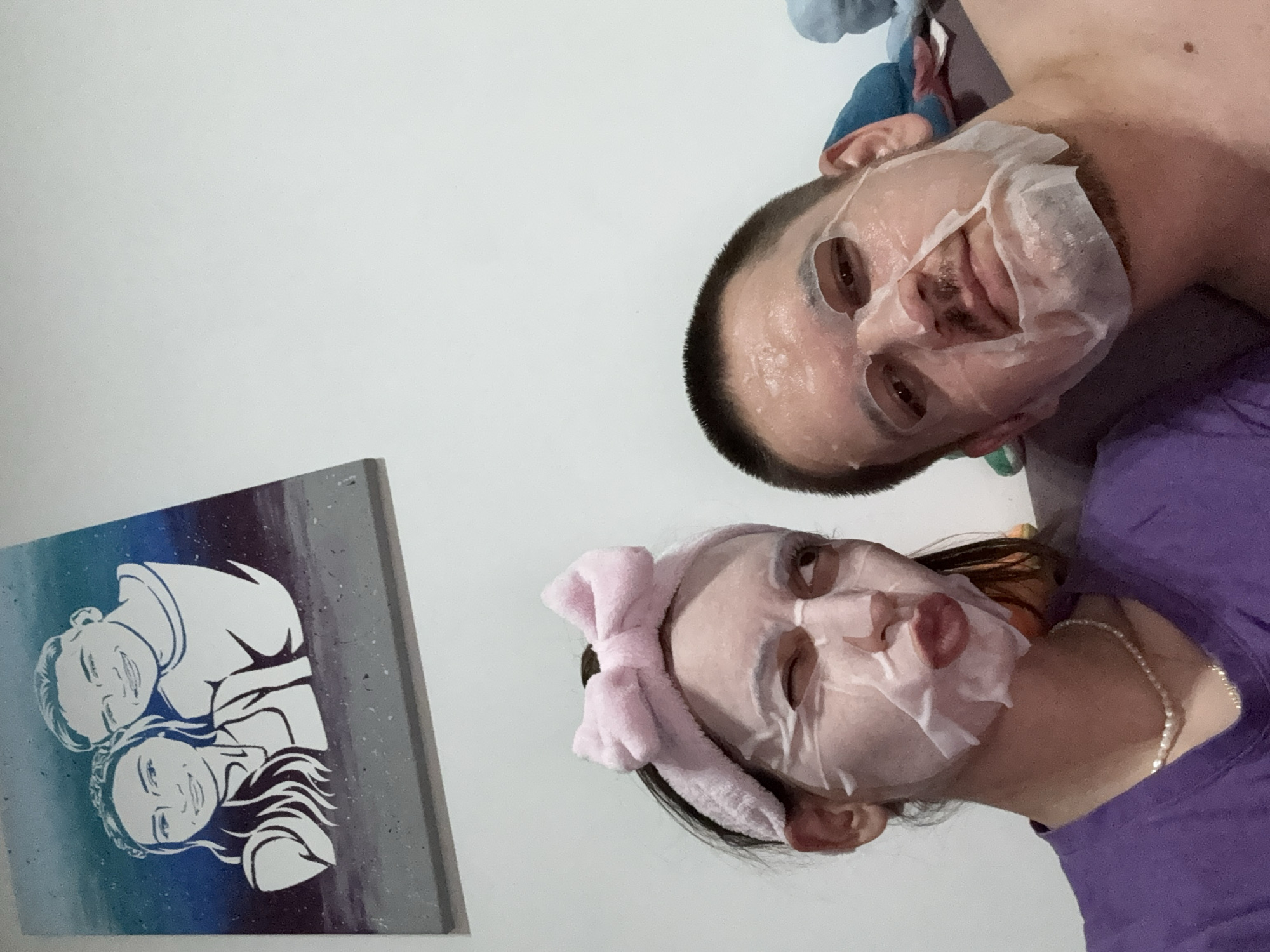 A selfie taken in an indoor setting shows a man and a woman wearing facial sheet masks. The man is positioned in the upper right of the frame, and the woman is in the lower right. The woman wears a pink headband. In the lower left corner, there is a painting of a couple. The background is a plain white wall. The image was not taken in Plovdiv, Bulgaria.