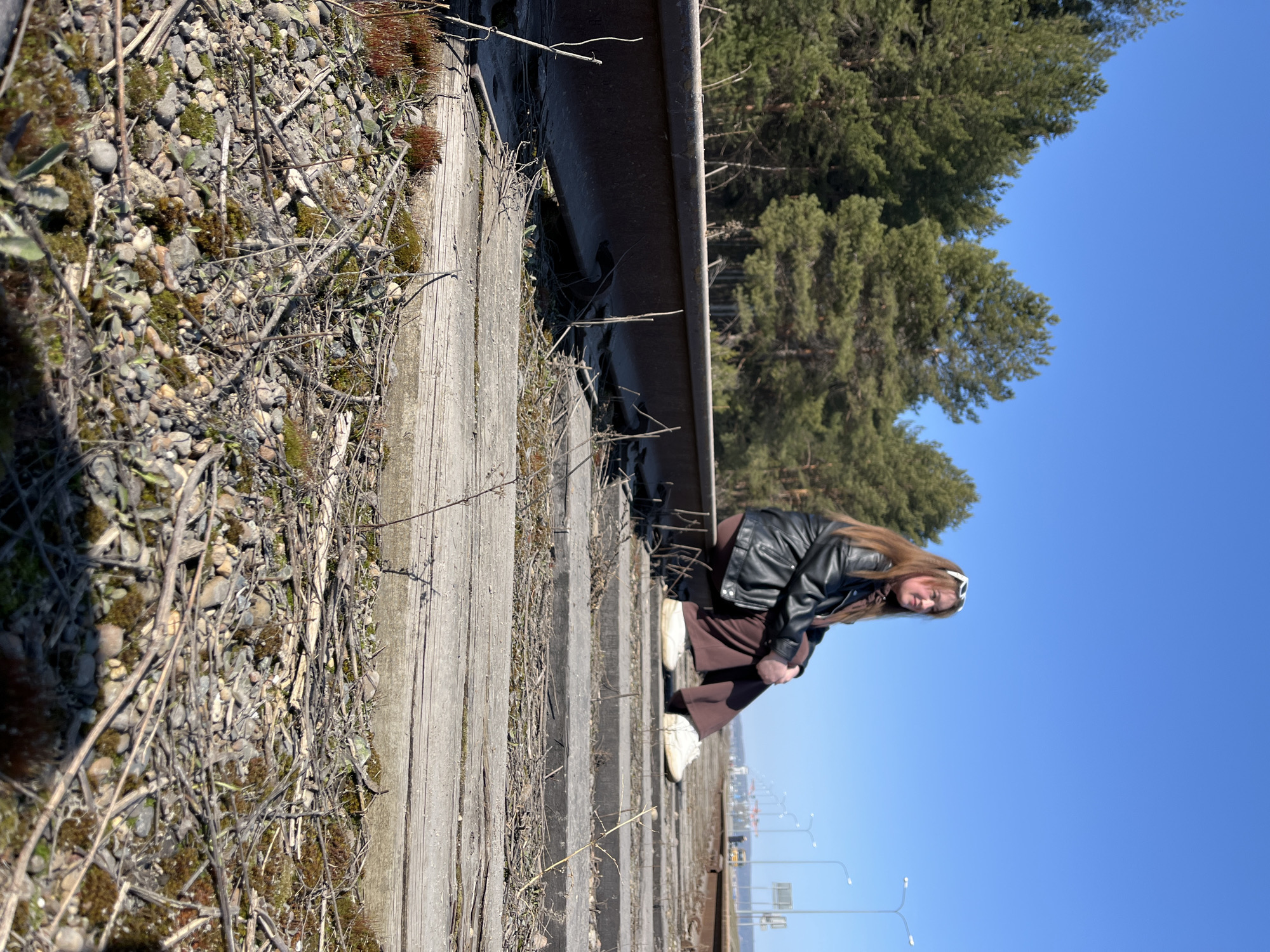 A woman with long, light brown hair wearing sunglasses, a black leather jacket, and brown trousers is squatting on train tracks, looking towards the camera. The train tracks are wooden and rusty. To the left of the tracks, there are rocks, moss, and dry twigs. In the background, there are pine trees and a bright blue sky. In the distance, there are light poles, buildings, and vehicles suggesting a cityscape. The location is likely Chaikovsky, Russia, due to its association with pine forests and urban development.