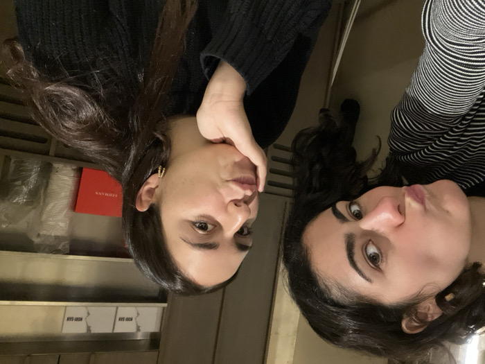 Two women pose closely together for a selfie. The woman on the left, wearing a black top, rests her hand on her cheek. Her dark hair is styled with earrings. To her left, are multiple stacks of white boxes, with one red Leimann box on top of them. The woman on the right has dark, curly hair and is wearing a black and white striped top. Both faces are upturned, as the photo is taken from above.