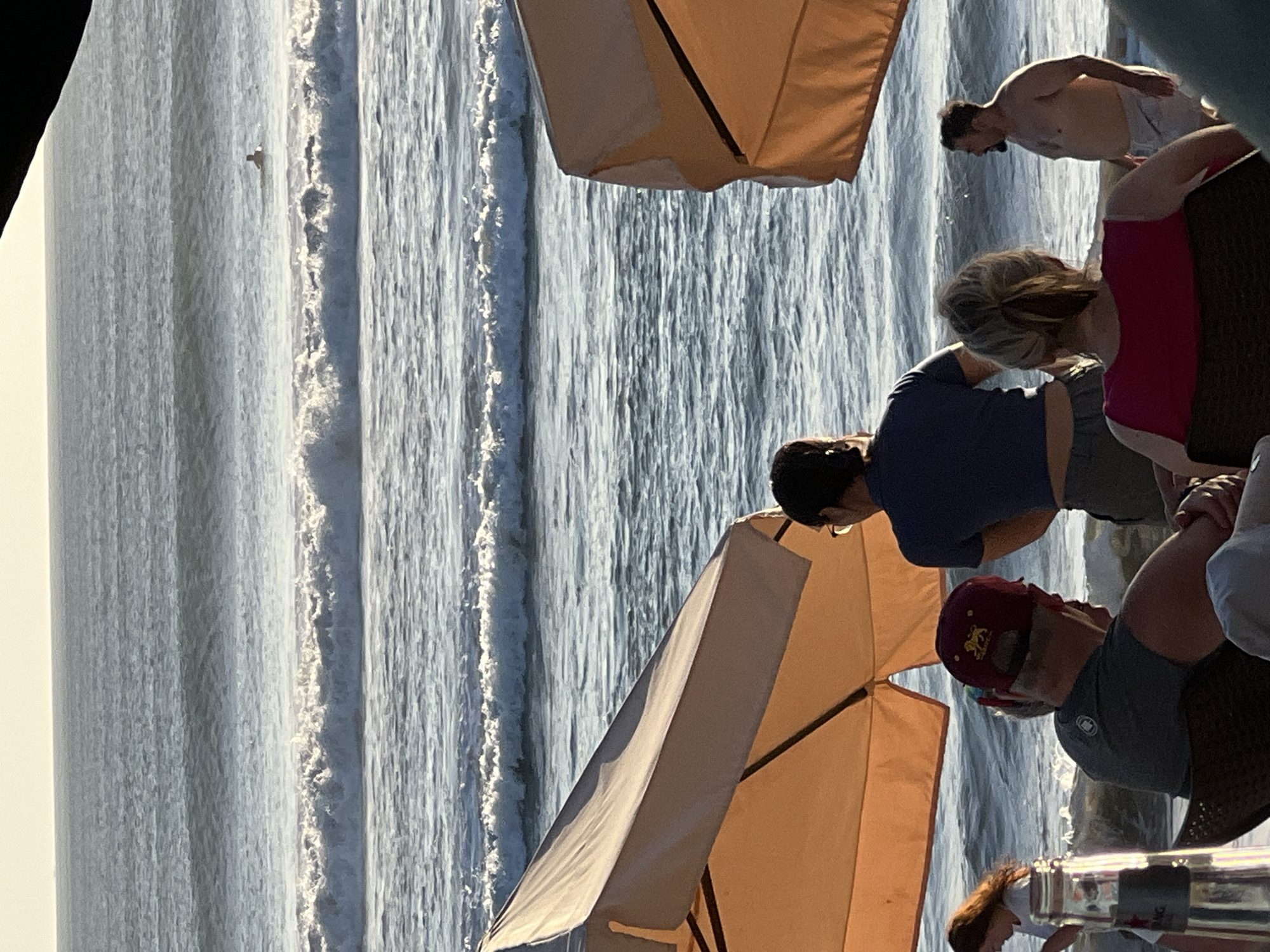 In Kuta, Indonesia, a group of people are seated at tables beneath large, light-colored umbrellas, facing the ocean. The water has significant waves, and there is one surfer visible in the distance. The sun is low in the sky, creating a warm glow over the scene. The foreground includes parts of the umbrellas, a bottle on a table, and several individuals seated in chairs. One person in the foreground is wearing a red baseball cap with a lion emblem. Another individual is standing and looking out at the ocean. The overall atmosphere appears to be relaxed and social, consistent with a beachside dining or lounge area.
