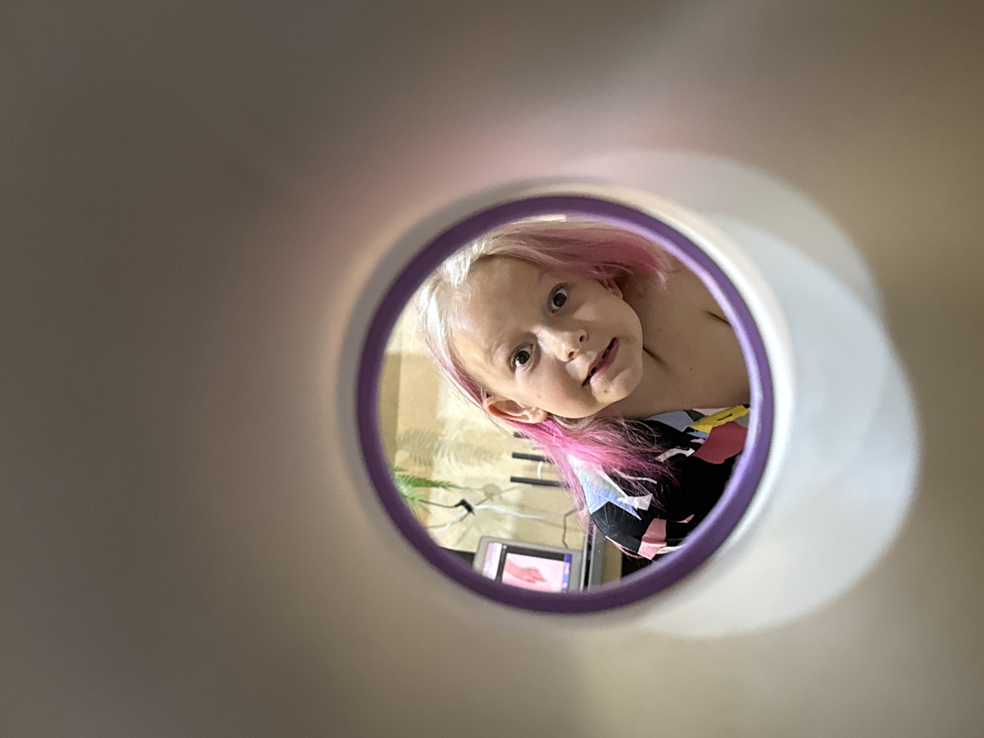 The image captures a close-up perspective of a young child looking into a circular opening, creating a unique framing. The photograph appears to be taken from inside a tube, pipe, or similar object, looking out at the child. This opening has a distinct purple rim.
The primary subject is a young girl with fair skin, light blonde hair, and noticeable pink streaks or dyed sections in her hair. She is looking directly into the camera with wide, curious eyes and a slightly tilted head. Her expression is observant and engaging. She is wearing a multi-colored garment featuring geometric patterns in black, white, pink, and blue, with a hint of yellow.
Through the circular frame, parts of an indoor background are visible. To the left and below the child's head, a screen or monitor is discernible, displaying what appears to be an image of a hand. Above the monitor, some shelving or furniture with horizontal lines can be seen, along with a partial view of a green plant. The overall setting suggests a domestic or playroom environment. No specific time of day or weather conditions can be determined, but the lighting suggests an interior during daylight hours. There is no visible text in the image.