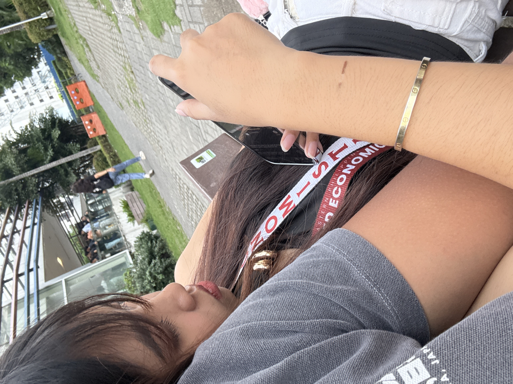 A medium-shot, low-angle, and eye-level shot shows a person lying down, holding a black smartphone. The individual has long, dark hair and is wearing a gray shirt and a headband with "PHILIPPINE EASTERN PETROLEUM CORPORATION" printed on it. A gold bracelet adorns their right wrist. In the background, figures can be seen walking near a building and some vegetation. Two orange cornhole setups are positioned near a pathway. The location is potentially within Davao City, Philippines, as indicated by the headband.
