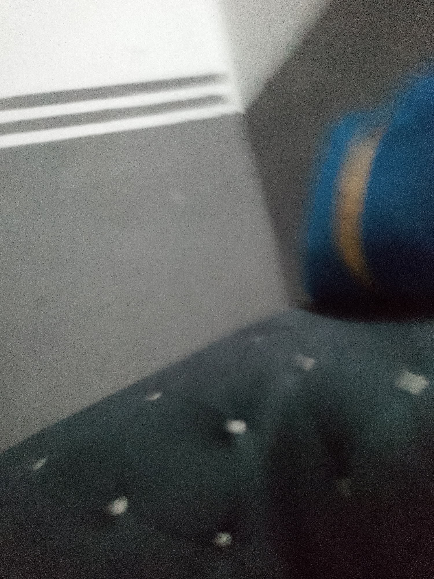 The image is a blurry, low-resolution photograph taken indoors, likely in a residential setting. The foreground shows a dark, textured surface, possibly a sofa or cushion, with several bright, rectangular shapes scattered across it, which could be reflections or light artifacts. In the background, a wall is visible, with a section of horizontal lines, suggesting a window or vent. To the right, a large, out-of-focus object with blue and yellow/orange coloring is partially visible, possibly a piece of furniture or a large toy. The overall impression is one of a dimly lit room with an abstract quality due to the blurriness and graininess of the image. There is no information in the image that can identify the location as Jauro Yinu, Nigeria, or any specific city.