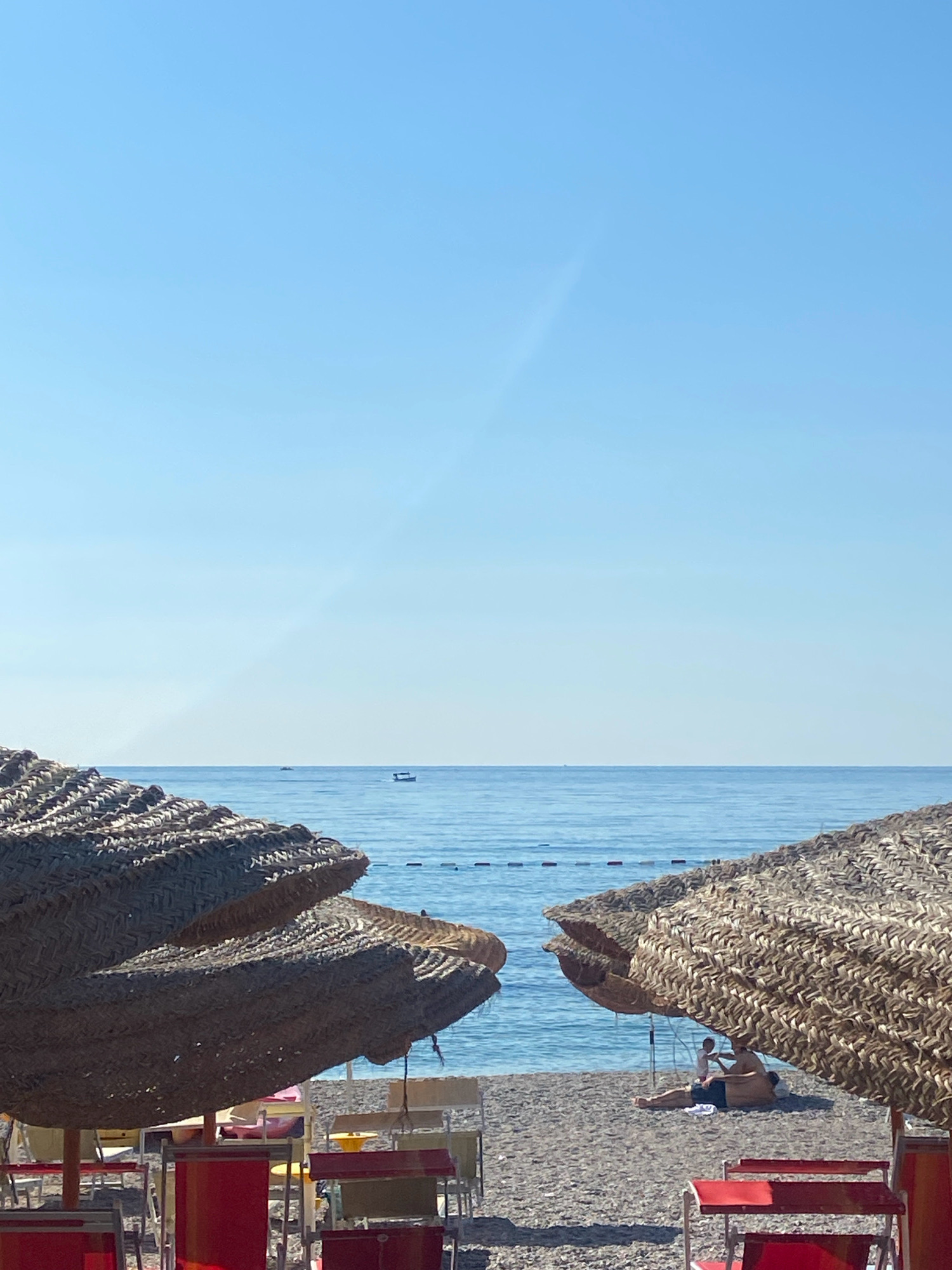The image depicts a serene beach scene under a clear blue sky. In the foreground, two large, thatched umbrellas are positioned on a pebble beach, angled towards the viewer. The umbrellas provide shade for several rows of empty red and yellow beach chairs and tables.
In the mid-ground, the calm, blue ocean stretches out to the horizon, where a faint line of boats is visible. A man and a child are seated under one of the umbrellas, appearing to be relaxing. The beach is sparsely populated, suggesting a peaceful atmosphere. The location is identified as Prijevor, Montenegro, a coastal town known for its beaches. The overall impression is one of a tranquil summer day by the sea.