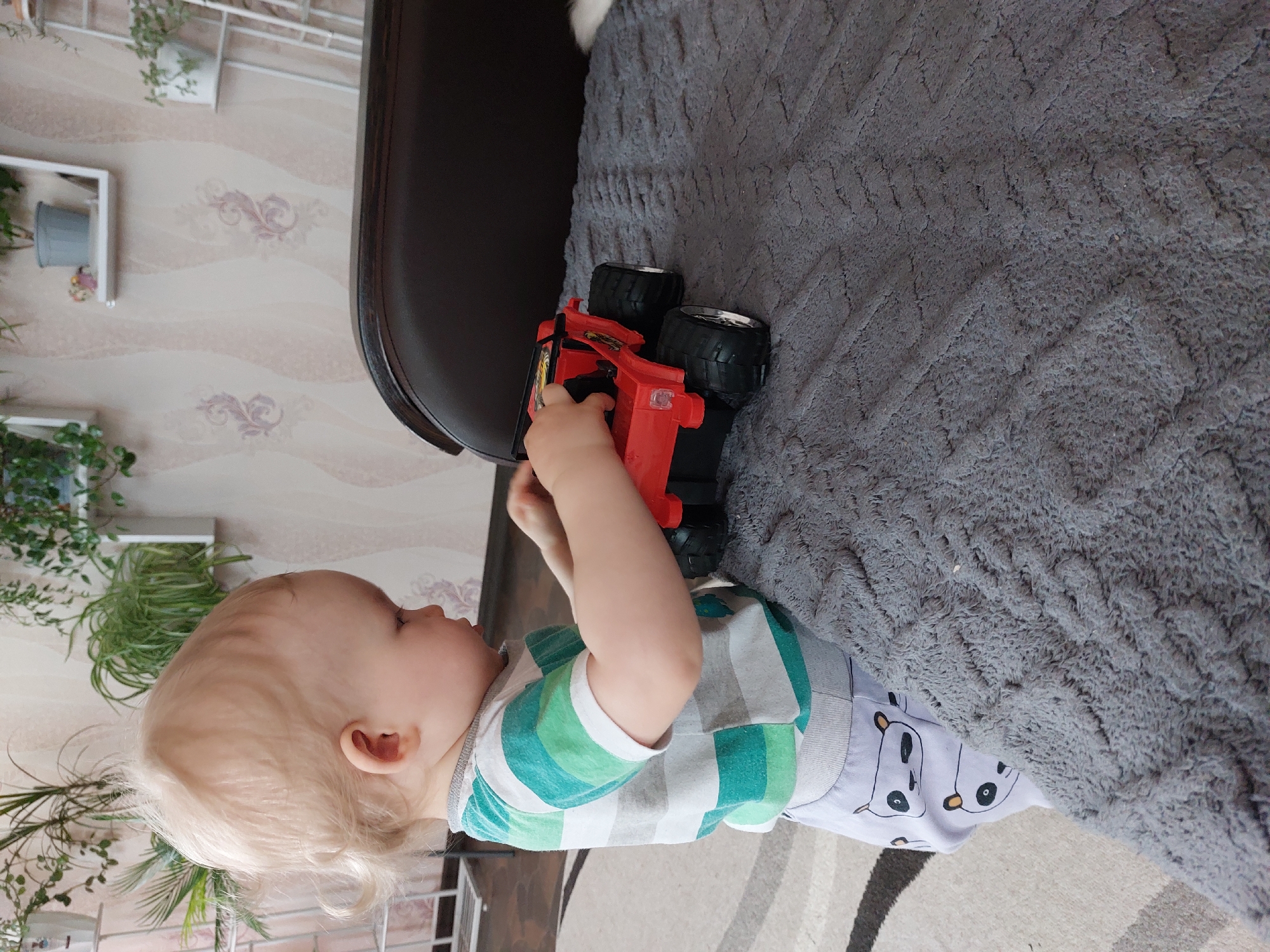 A small child with blond hair is playing with a red toy truck on a gray blanket. The child is wearing a striped green and white shirt and light gray pants with a panda pattern. A brown couch is next to the blanket. In the background is a wall with shelves holding plants. The location is Большой Букор, Russia.
