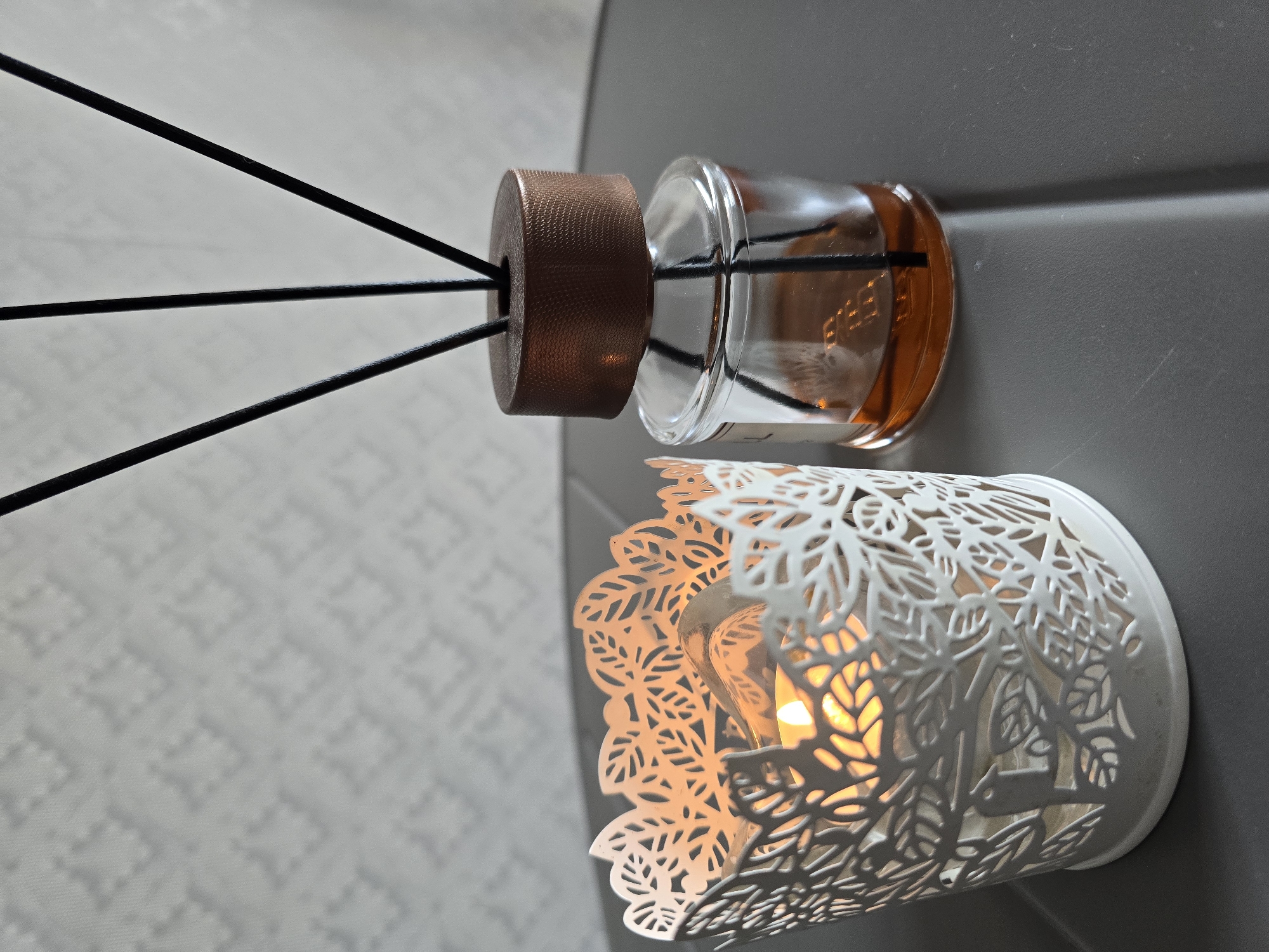 In Sofiivska Borshchahivka, Ukraine, on a dark grey, textured surface, two decorative objects are placed side-by-side. On the left, a white, filigree metal candle holder with a leaf motif encloses a lit candle, casting a warm orange glow. The candle holder appears to have a cutout of a bird within its design. To the right of the candle holder is a clear glass diffuser bottle containing amber-colored liquid and topped with a textured brown cap. Several black reeds extend upwards from the diffuser. The background features a textured light grey wall with a subtle geometric pattern.