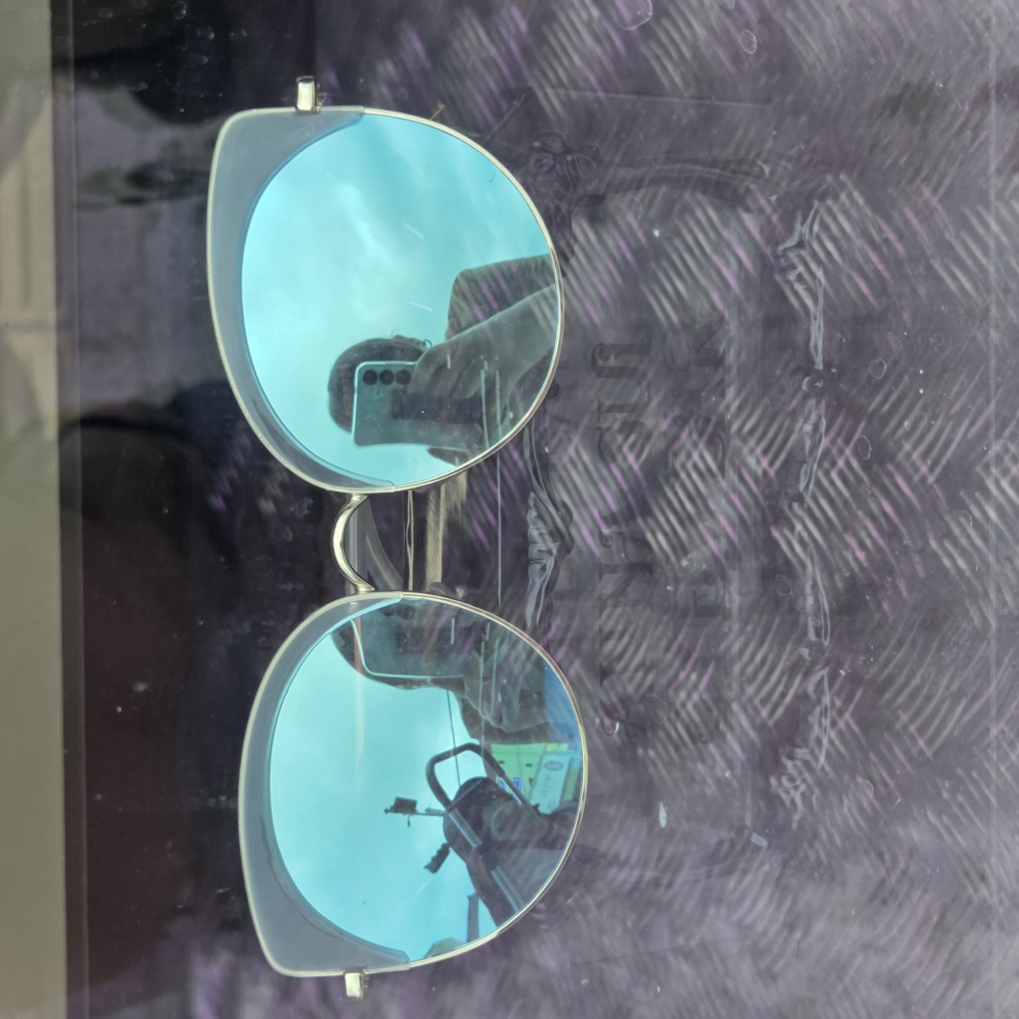 In an outdoor setting in Dunnae-myeon, South Korea, a pair of blue-tinted sunglasses with a gold frame are the central subject. The sunglasses are positioned on a surface that appears to be a metallic floor or walkway with a diamond plate pattern, featuring streaks of purple and white. Reflections in the lenses reveal a person holding a smartphone, taking a selfie, with a blurred background of what seems to be a street scene, including a motorcycle, a pole with a sign, and some buildings. The overall lighting suggests it is daytime.