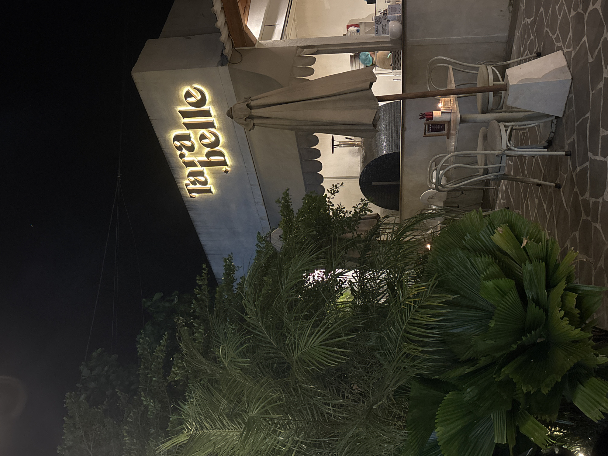 The image captures the exterior of "Tarabelle" in Ungasan, Indonesia, at night. The restaurant's illuminated sign is prominently displayed on the side of a building. An off-white patio umbrella and several white chairs and tables are visible outside. Dense foliage partially obscures the view. The pavement is paved with irregular stones. The night sky is black.