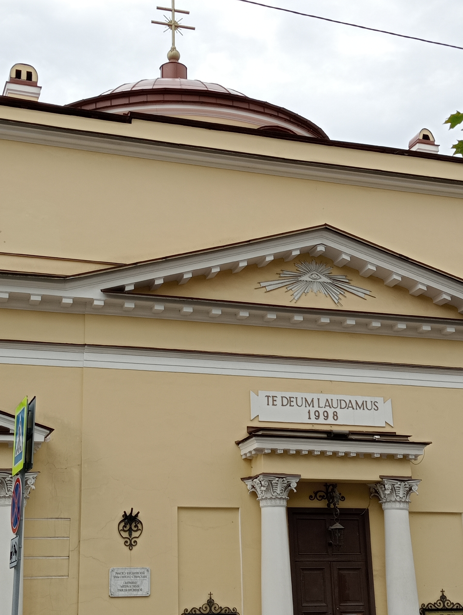 The image shows a facade of a light yellow building, likely a church, in Saint Petersburg, Russia. The building has a prominent reddish-brown dome topped with a cross. There is a decorative pediment above a central entrance, featuring an all-seeing eye symbol within a radiating sunburst design. Below the pediment, a plaque reads "TE DEUM LAUDAMUS 1998". The entrance itself is a dark wooden door flanked by two white classical columns with ornate capitals. To the left of the entrance, a historical plaque and a decorative wrought-iron element are visible on the wall, along with a street signpost. The sky is overcast, suggesting a cloudy day.
