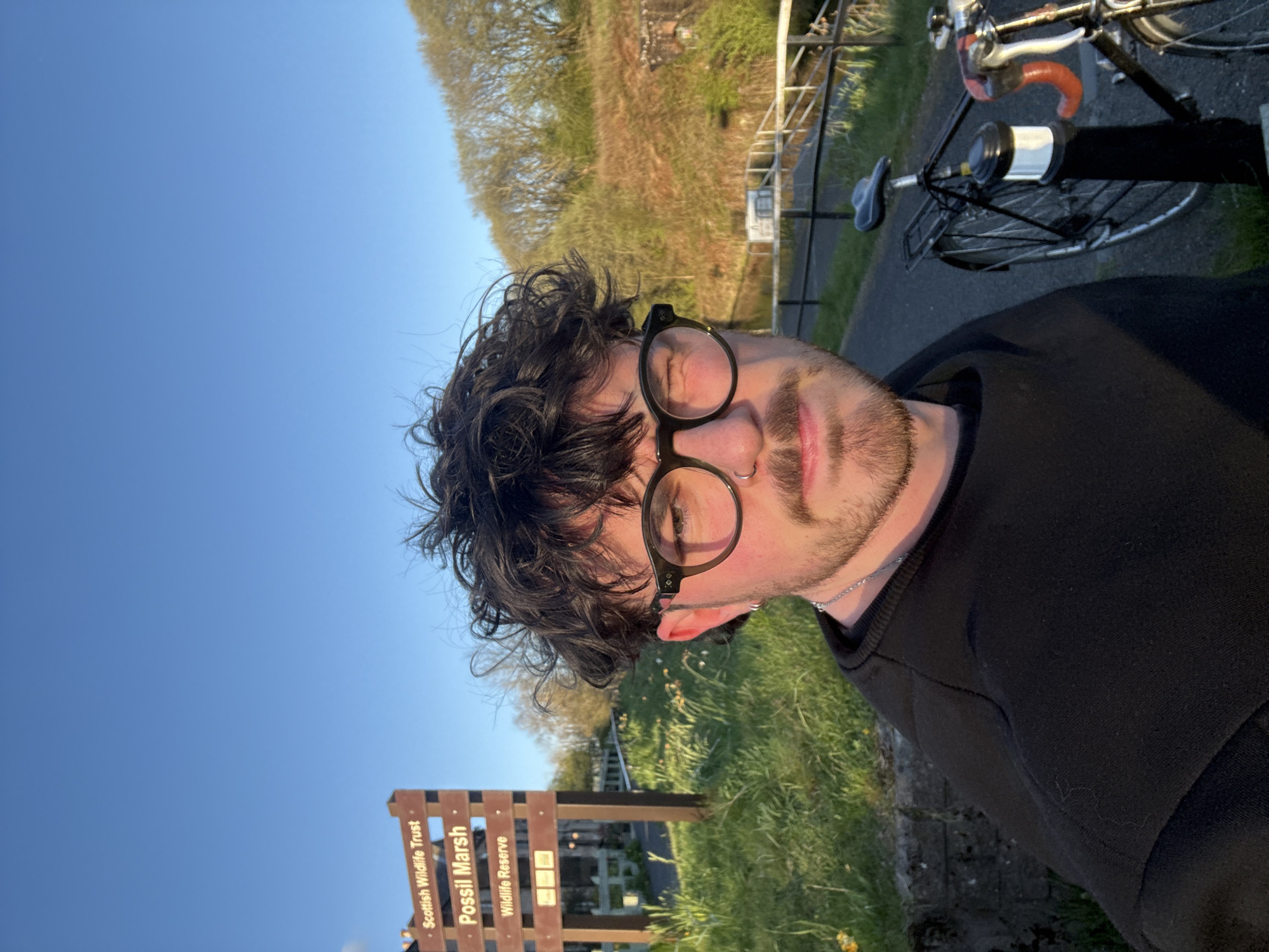 The image features a person in a selfie-style shot, oriented sideways in the original, but depicting a clear, sunny outdoor scene. The subject is a person with dark, curly hair, wearing circular dark-rimmed glasses, a nose ring, and a neatly trimmed beard or stubble. They are wearing a dark long-sleeved top and a silver chain around their neck, looking directly at the camera with a neutral to slightly smiling expression.
The setting appears to be a nature reserve or park. In the background, to the right of the person, a bicycle is partially visible on what looks like a paved path or road. Lush green grass and distant trees or bushes line the background, illuminated by warm, directional sunlight, suggesting late afternoon or early evening (golden hour). The sky above is clear and blue.
In the bottom left corner, a wooden sign is prominently displayed. The text on the sign clearly reads:
* "Scottish Wildlife Trust"
* "Possil Marsh"
* "Wildlife Reserve"
This identifies the location as the Possil Marsh Wildlife Reserve, managed by the Scottish Wildlife Trust. The overall atmosphere is peaceful and serene, capturing a moment outdoors.