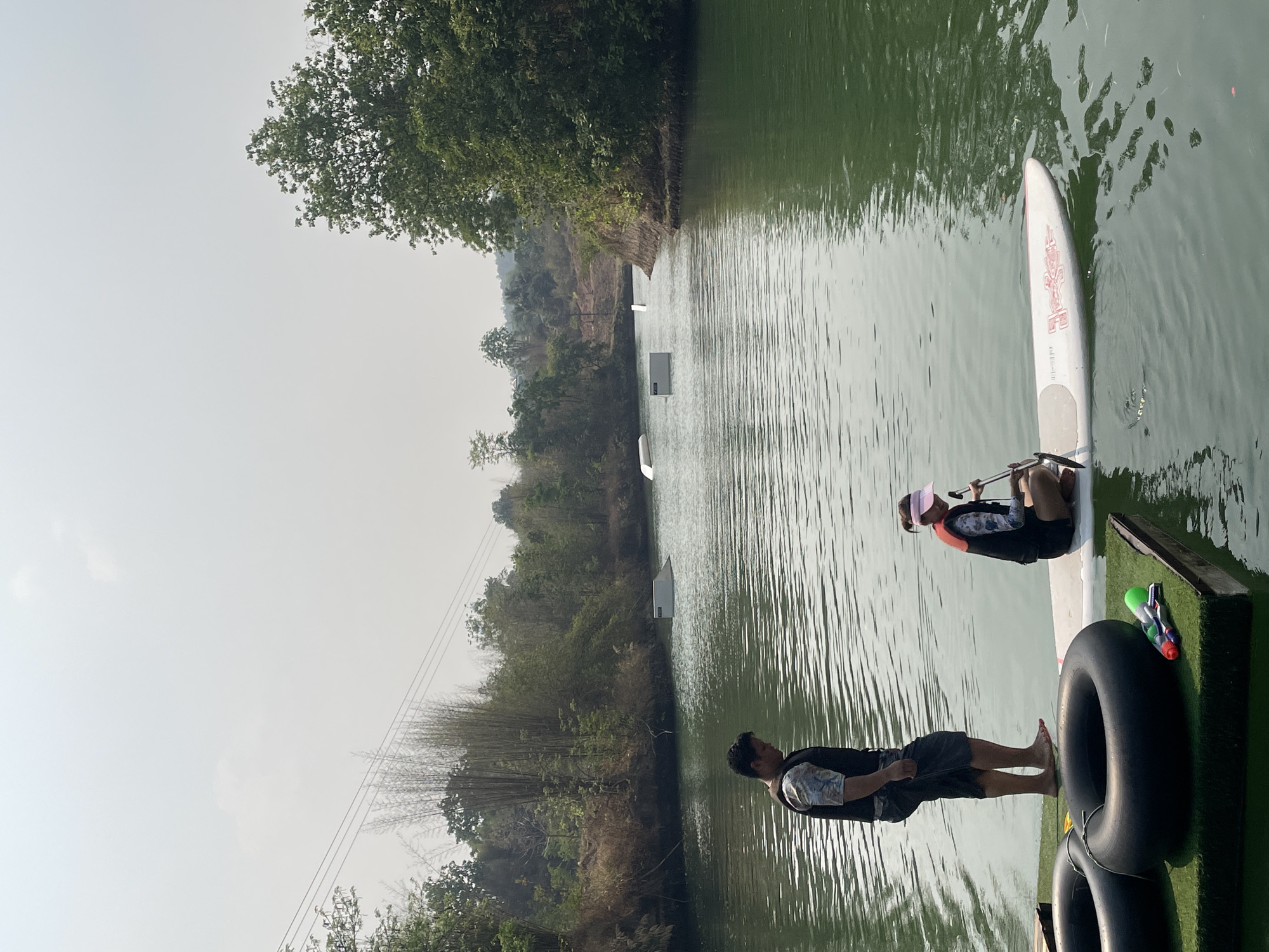 The image shows a scene on a body of water, likely a lake or river, in Tambon Nam Phrae, Thailand. The water is greenish-brown and calm, with light reflecting off the surface. There are trees and vegetation along the banks of the water. In the distance, there are wakeboarding obstacles set up on the water.
Two people are visible on a floating platform. One person is sitting on a paddleboard, wearing a life vest and a visor. They appear to be preparing to paddle. The other person is standing on the platform, dressed in shorts and a shirt, looking at the person on the paddleboard. Two black inner tubes and a water gun are on the platform. Powerlines can be seen in the background.