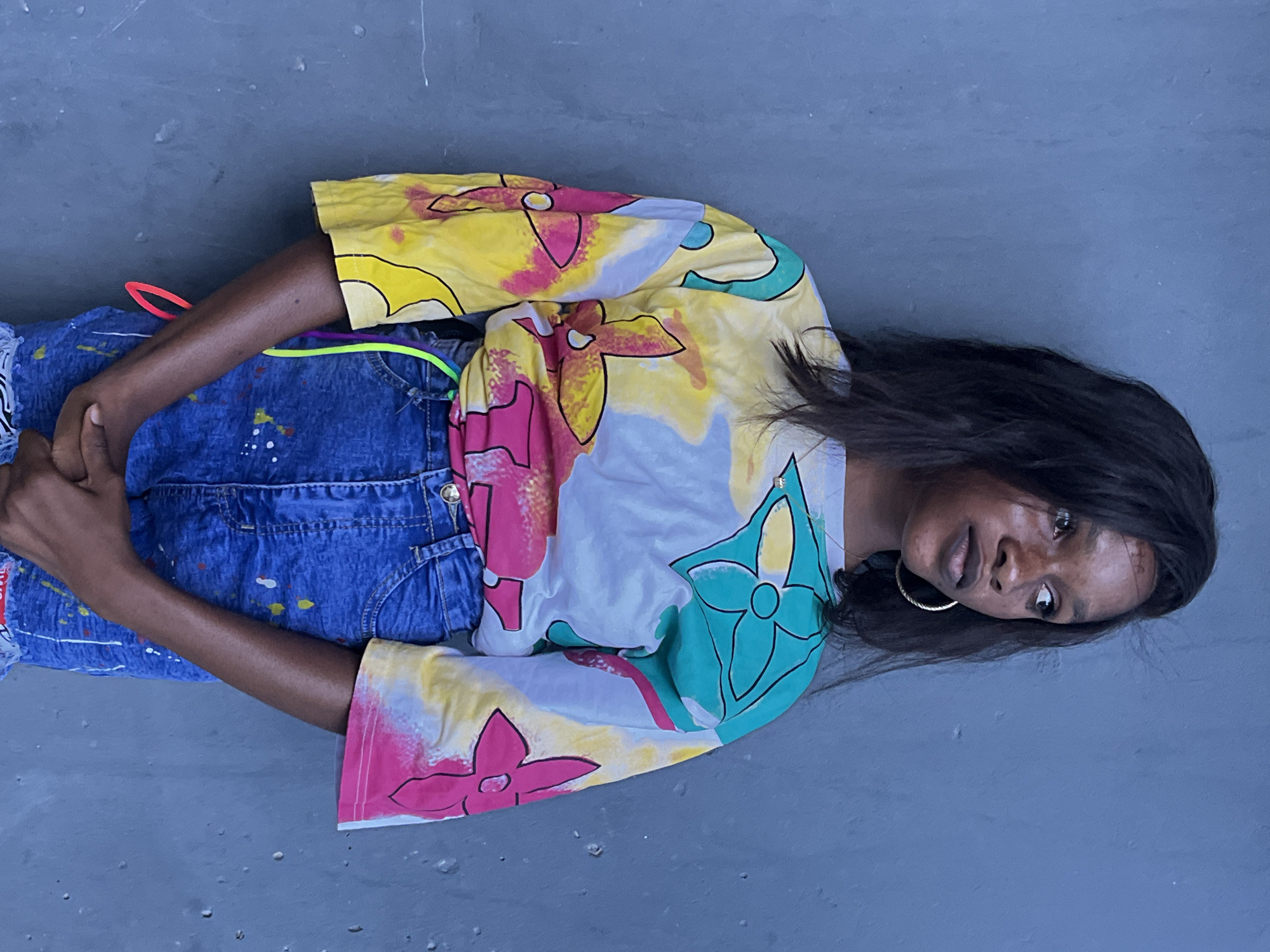 A young Black woman is lying on a gray surface, possibly concrete. She is wearing blue denim overalls with paint splatters and a colorful patterned shirt with yellow, pink, and green floral designs. The shirt appears to be tie-dyed. She has long dark hair and is wearing a gold hoop earring. The location is likely Badagry, Nigeria, although there are no specific background elements to confirm this. The woman is looking towards the camera with a neutral expression. Her hands are clasped together. There is an orange and a green neon accessory near her waist.