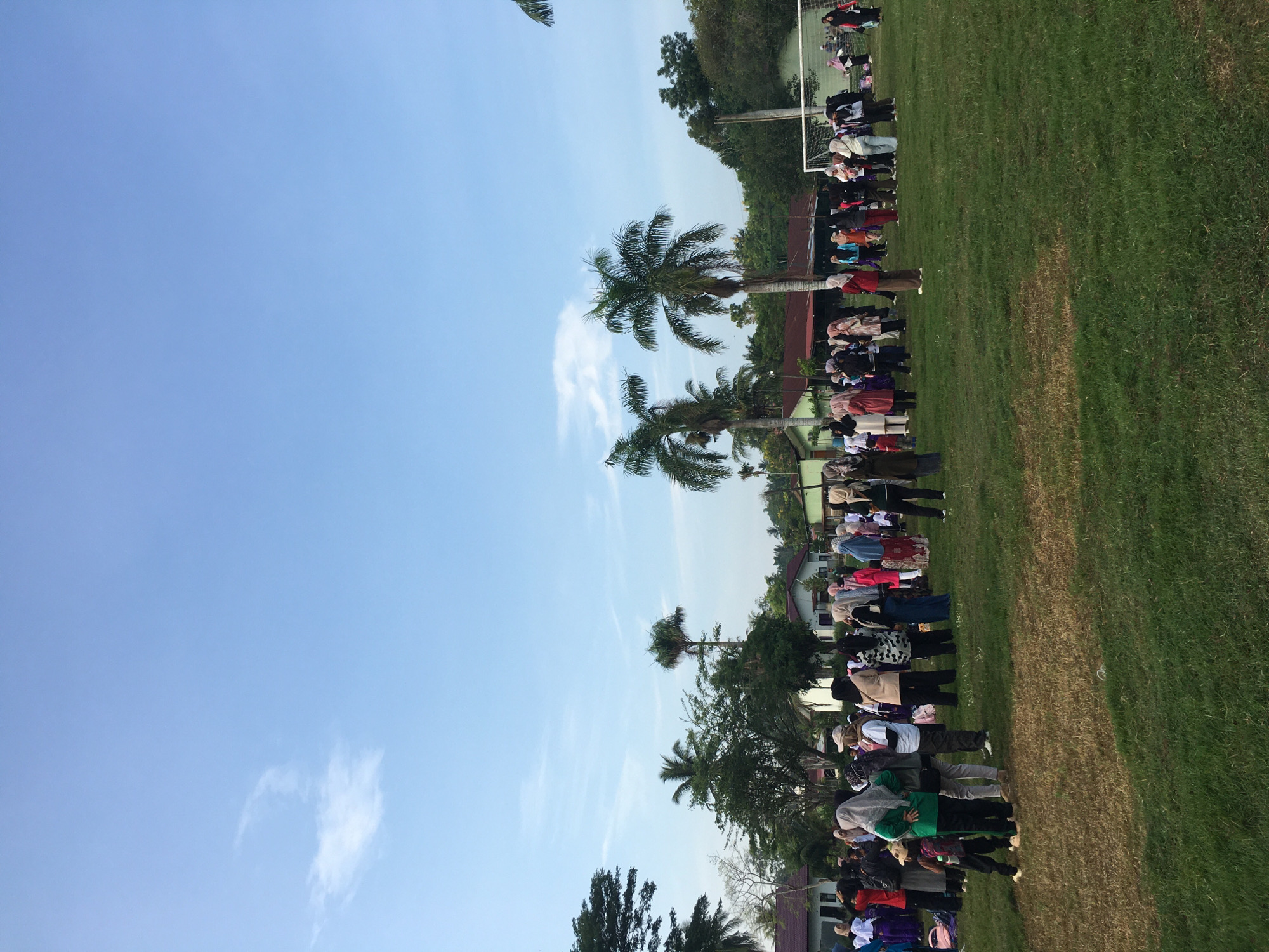 A group of people stand in neat rows on a grassy field. The location is likely a schoolyard in Lhokseumawe, Indonesia. A blue sky with scattered clouds is overhead. Palm trees line the field. The people in the rows are all facing the same direction. The purpose of the gathering is not clear, but it may be a formal ceremony or a communal prayer.