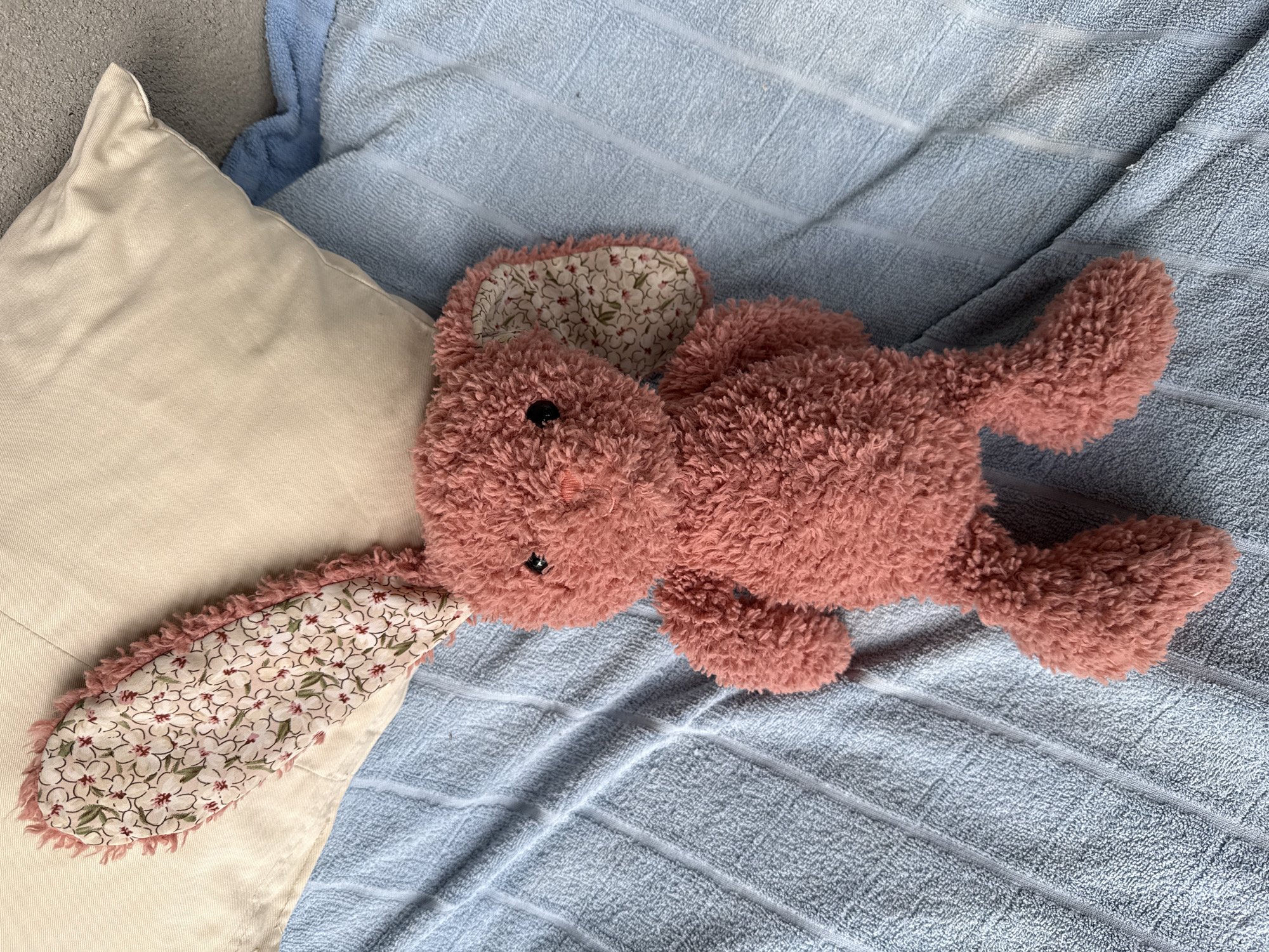 A plush pink bunny toy lies on a textured blue towel. The bunny's ears are lined with a floral fabric. Behind the bunny and towel, a light-colored pillow rests against a gray surface. The scene is indoors, with no indication of Dilijan, Armenia.