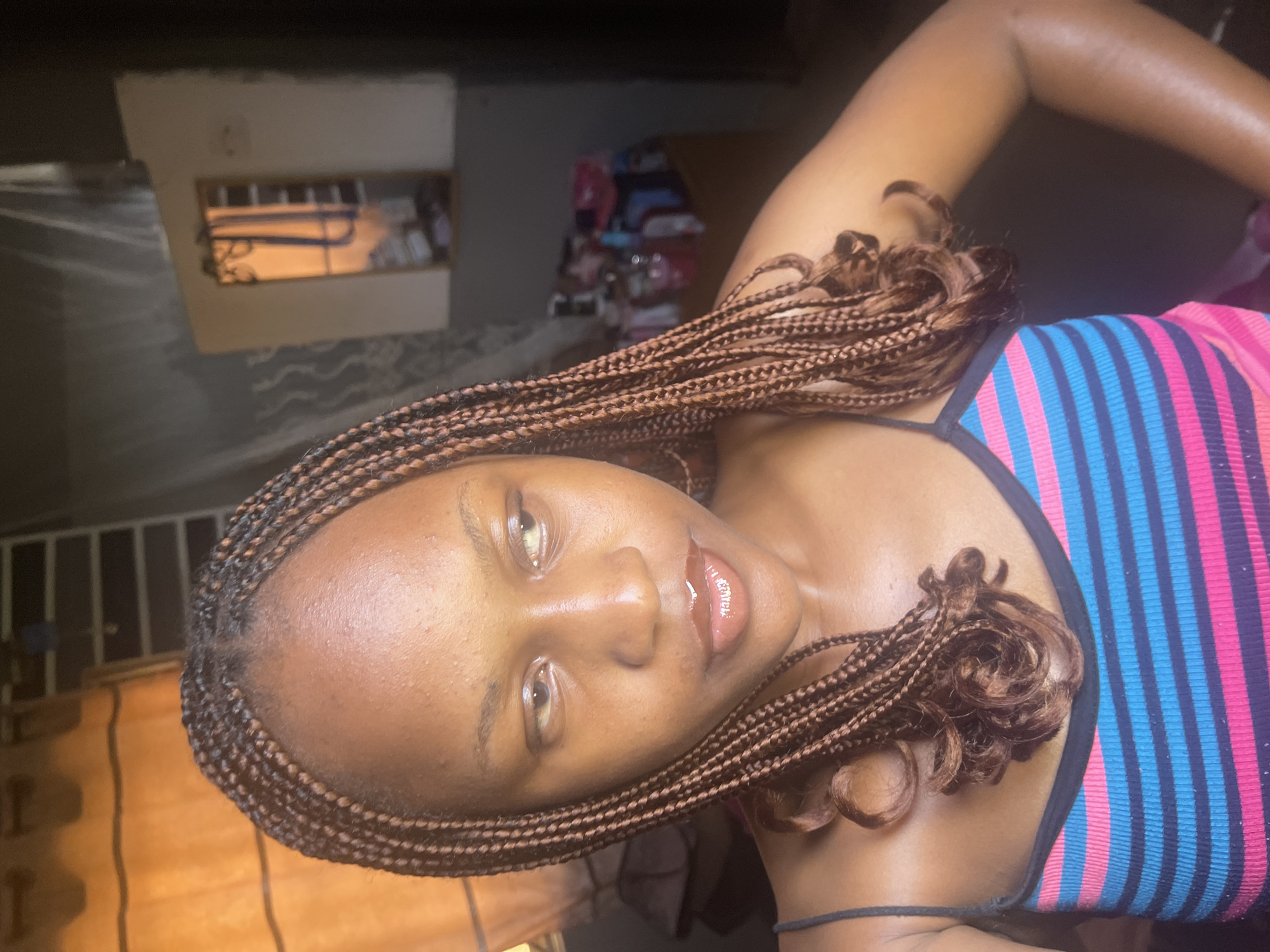 This media file is a close-up, portrait-style photograph of a young Black woman. She has her hair styled in numerous thin braids, with some ends curled. She is wearing a tank top with horizontal stripes in pink, blue, and black. Her head is tilted, and she is looking slightly off-camera with a subtle smile. Her lips are glossy.
In the background, there are some indistinct objects and textures. To the left, there is a shelf or box-like structure with items visible inside, illuminated by a warm light. Further in the background, there are stacked items, possibly clothing or fabric, with colorful patterns. The overall lighting suggests an indoor setting, possibly at dusk or night. The mood of the image is relaxed and candid.
There is no visible text in the image. The location context is provided as Rafin Par, Nigeria.