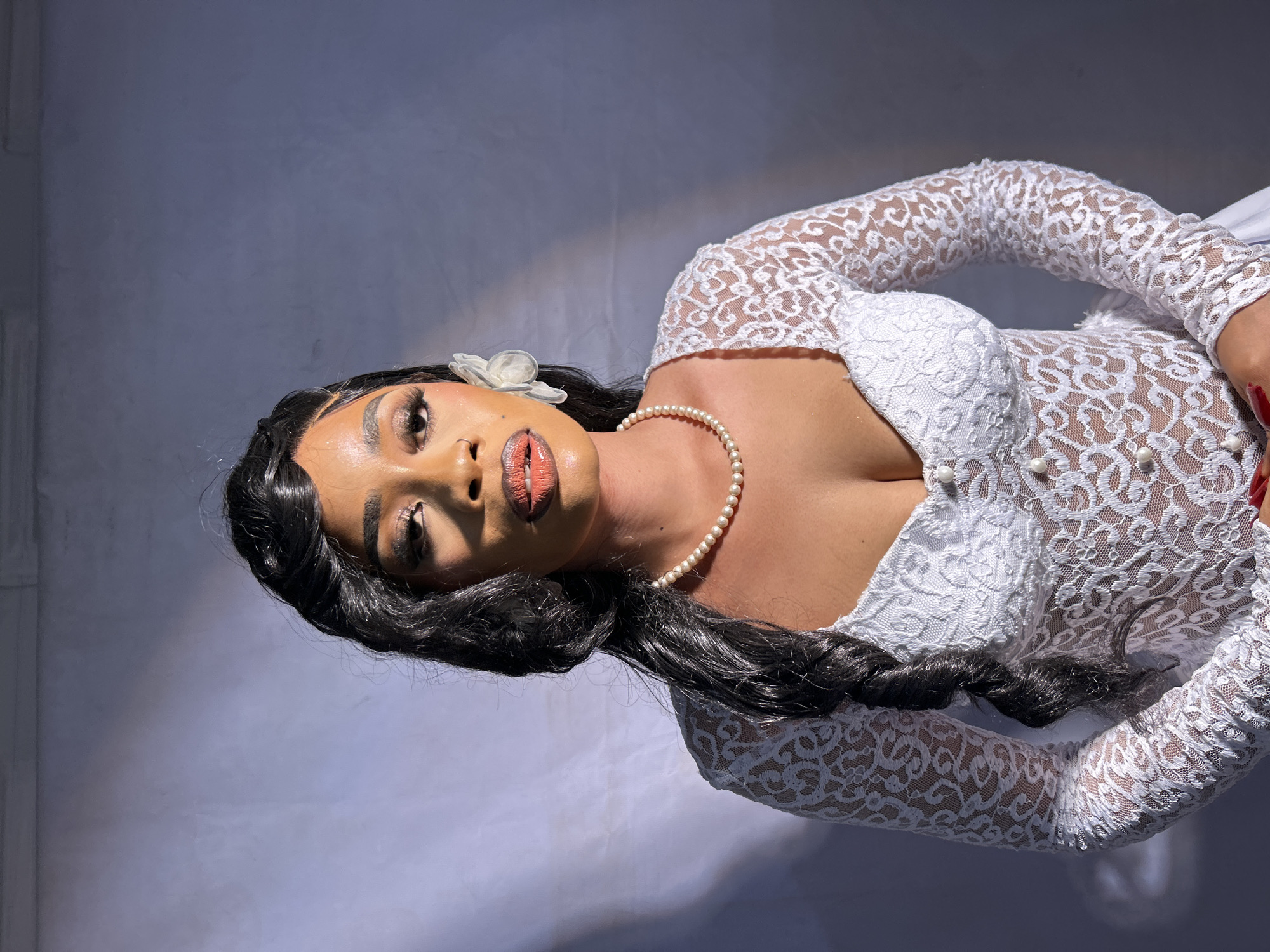 A woman with dark hair and fair skin is lying on a light gray background. She is wearing a white, lace dress with long sleeves and a sweetheart neckline. She also has a pearl necklace and a white flower in her hair. Her makeup includes frosted eyebrows and orange lipstick. The location is unknown and not in Jos, Nigeria.