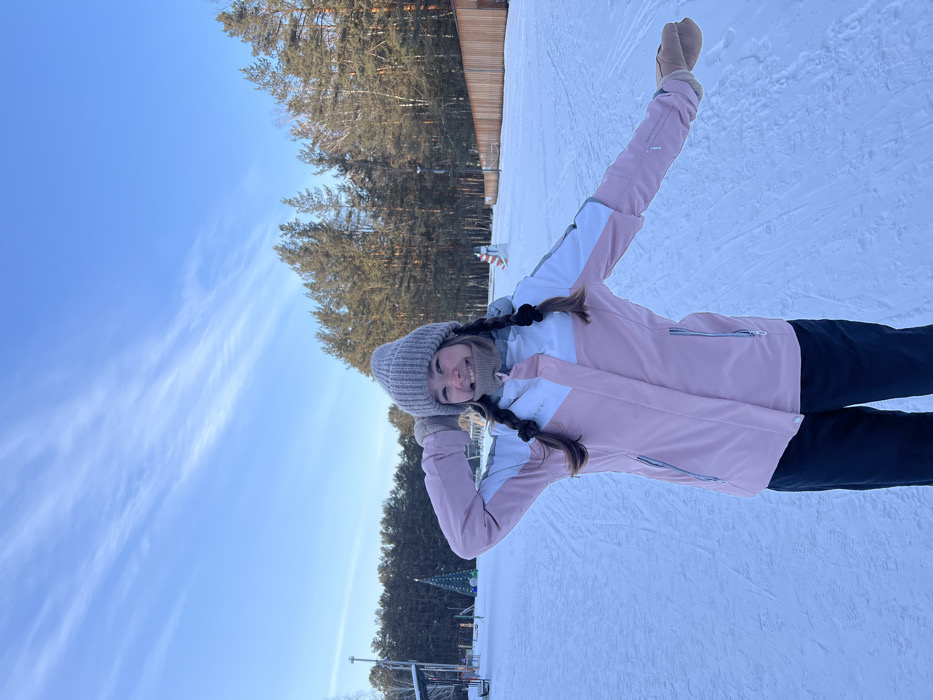 A young woman in winter clothing stands on a snowy surface, seemingly a ski slope. She wears a pink and white jacket, a gray knit hat, and tan mittens. Her hair is braided into two pigtails. She poses with her arms raised. Behind her is a clear blue sky with wispy clouds and a forest of tall pine trees. A wooden building is also visible in the background. The photo appears to be taken on a sunny day.