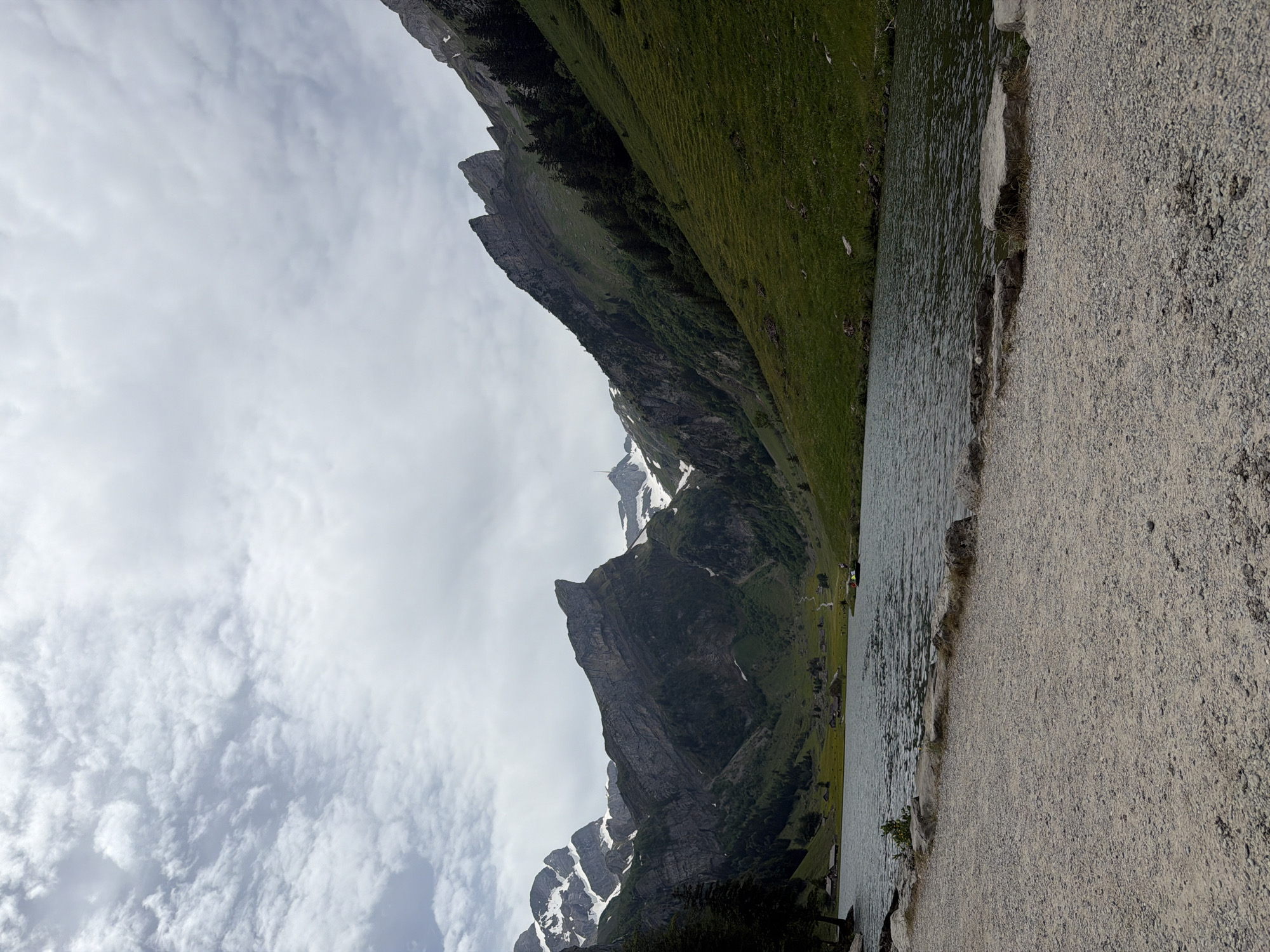 The image depicts a landscape scene at Seealpsee Lake in Schwende-Rüte, Switzerland. The view is primarily of the lake's edge, with a gravelly shoreline on the right side of the image. The water stretches towards the left, where steep, green slopes rise into imposing mountains. There's evidence of snow on some of the higher peaks. The sky is overcast with a variety of cloud formations. There appear to be some small buildings and trees along the far side of the lake, but people are not clearly visible.