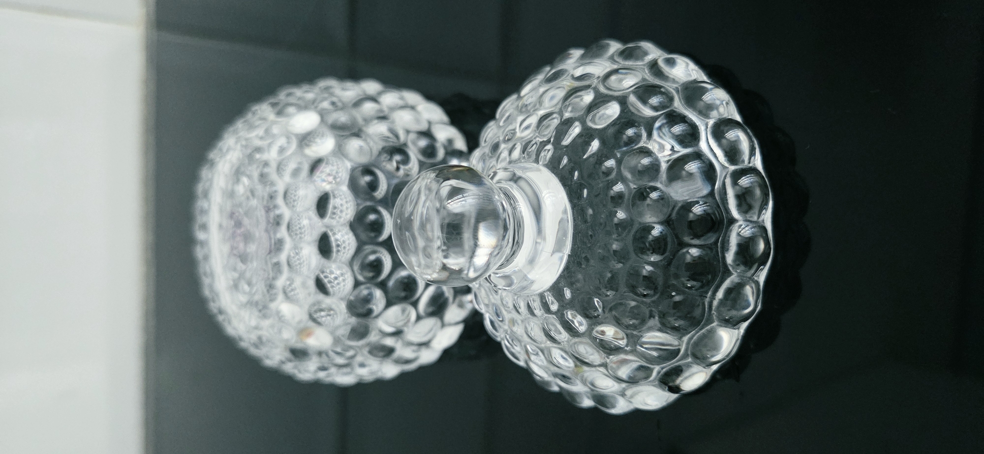 The image shows two clear glass bowls with a hobnail pattern, one nested inside the other. The front bowl has a round, clear glass knob on top of its lid, which is slightly tilted. The background bowl is less in focus and appears to be the base of a container. The objects are placed on a dark, reflective surface, possibly a table, with a lighter, blurred background. There are no people or animals visible in the image. The image does not provide any information about its location in the city of Lipetsk, Russia.