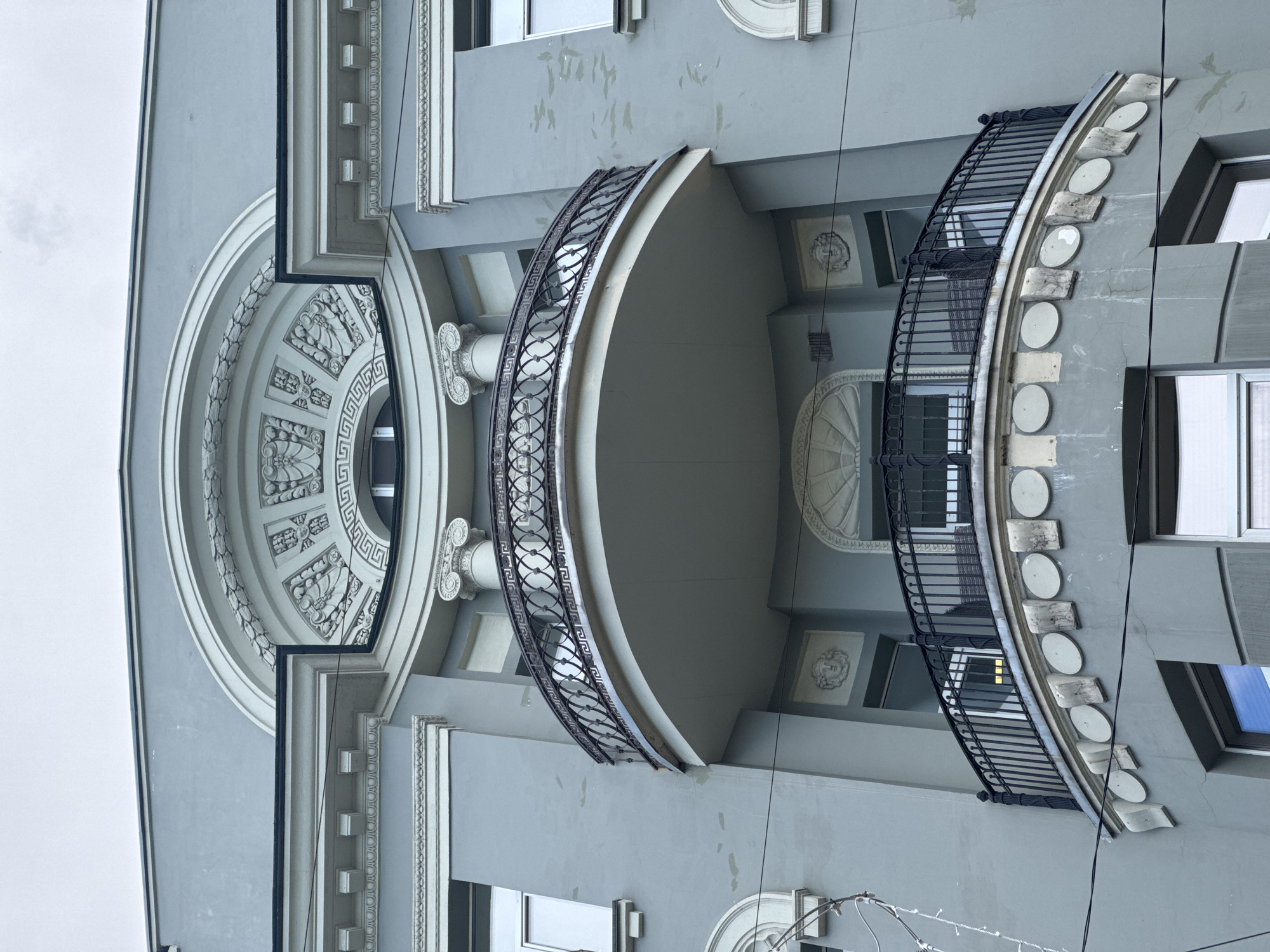 The image shows the facade of a building in Moscow, Russia. The architecture features classical elements, including a large circular window with decorative molding, columns, and two curved balconies with wrought iron railings. The building is painted in a light gray color, and the overall style suggests a historic structure. Electrical wires are visible against the backdrop of a cloudy sky.