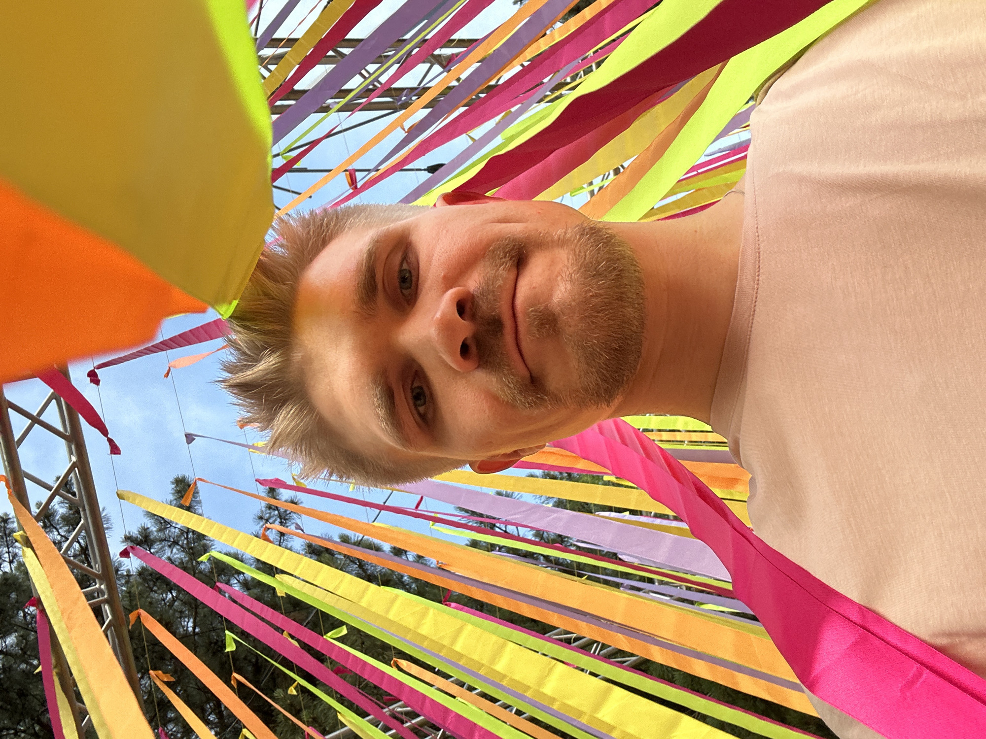 A man is looking down at the camera with a slight smile. He has light-colored hair and a trimmed beard. He is wearing a light-colored t-shirt. Above and around him, there are numerous brightly colored ribbons strung up. They are shades of orange, yellow, pink, and purple. In the background, some trees and a metal structure can be seen. The man's face and shirt are the primary subjects, with the ribbons forming a colorful, blurred background. The location is Tashkent, Uzbekistan.