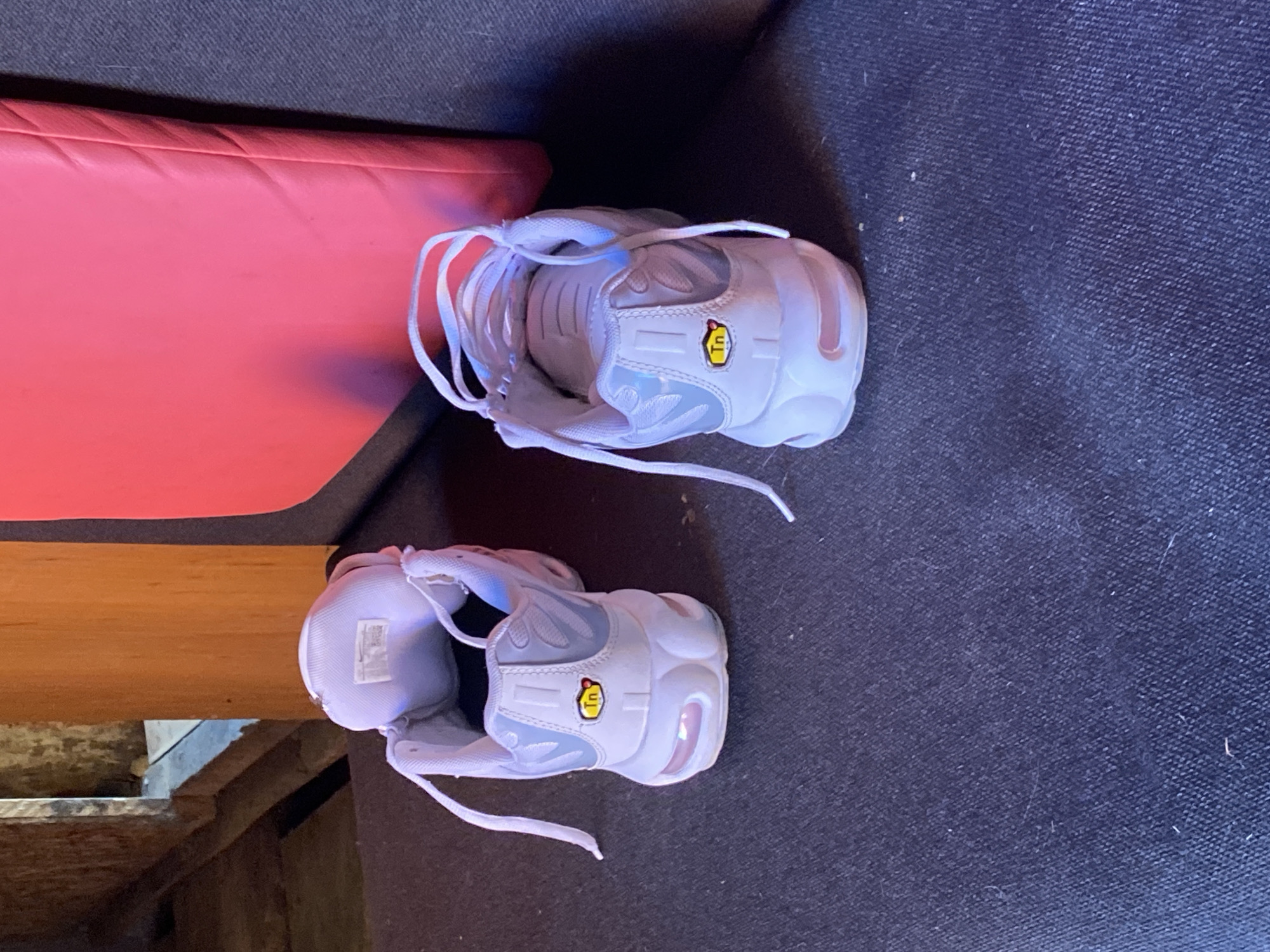 In Borodulino, Russia, a pair of white Nike Air Max Plus TN sneakers are placed on a dark blue, textured surface, likely upholstery. The shoes are positioned with their backs facing the camera, and their laces are loosely tied. A bright pink, rectangular object is visible in the background, partially obscuring a wooden structure. The lighting suggests an indoor environment.