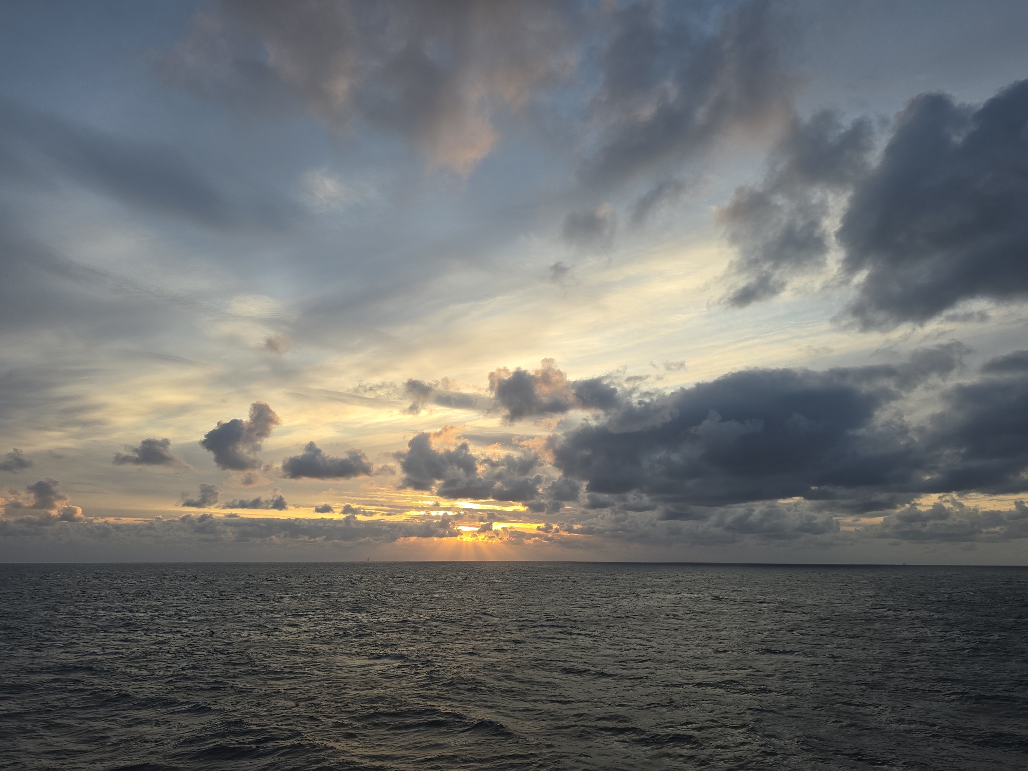 The image shows a body of water, possibly an ocean, under a sky filled with clouds. The sun is setting or rising on the horizon, casting a warm, orange light through the clouds. The clouds are a mix of light and dark shades, with some hints of pink and purple. The water is dark, reflecting the colors of the sky. The location is unknown.
