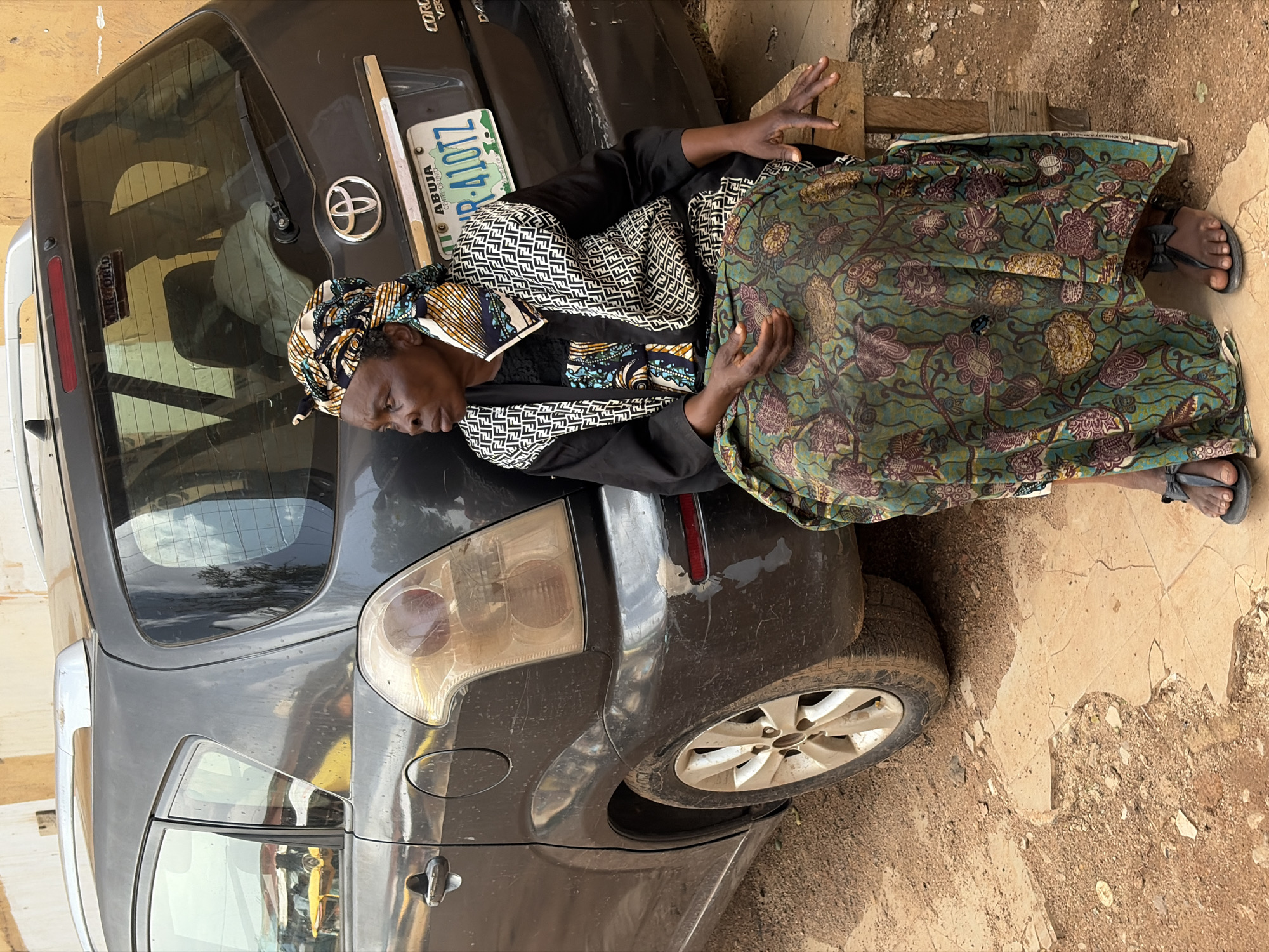 An older woman with dark skin and a headscarf sits on a wooden stool next to a dark grey Toyota minivan parked on a dirt and cracked concrete surface. The woman is wearing a patterned headscarf and a dark top with a black and white patterned overlay. Her skirt is long and green with a floral pattern. Her feet are bare, except for black flip-flops. The minivan is dirty, with dust on its exterior. The license plate is visible and reads "ABUJA CENTRE OF UNITY" and "NR 410TZ." Behind the woman, the rear of the minivan is visible, including a wiper blade and a reflection of a building in the window. The environment appears to be an outdoor urban setting, possibly a roadside or a yard.