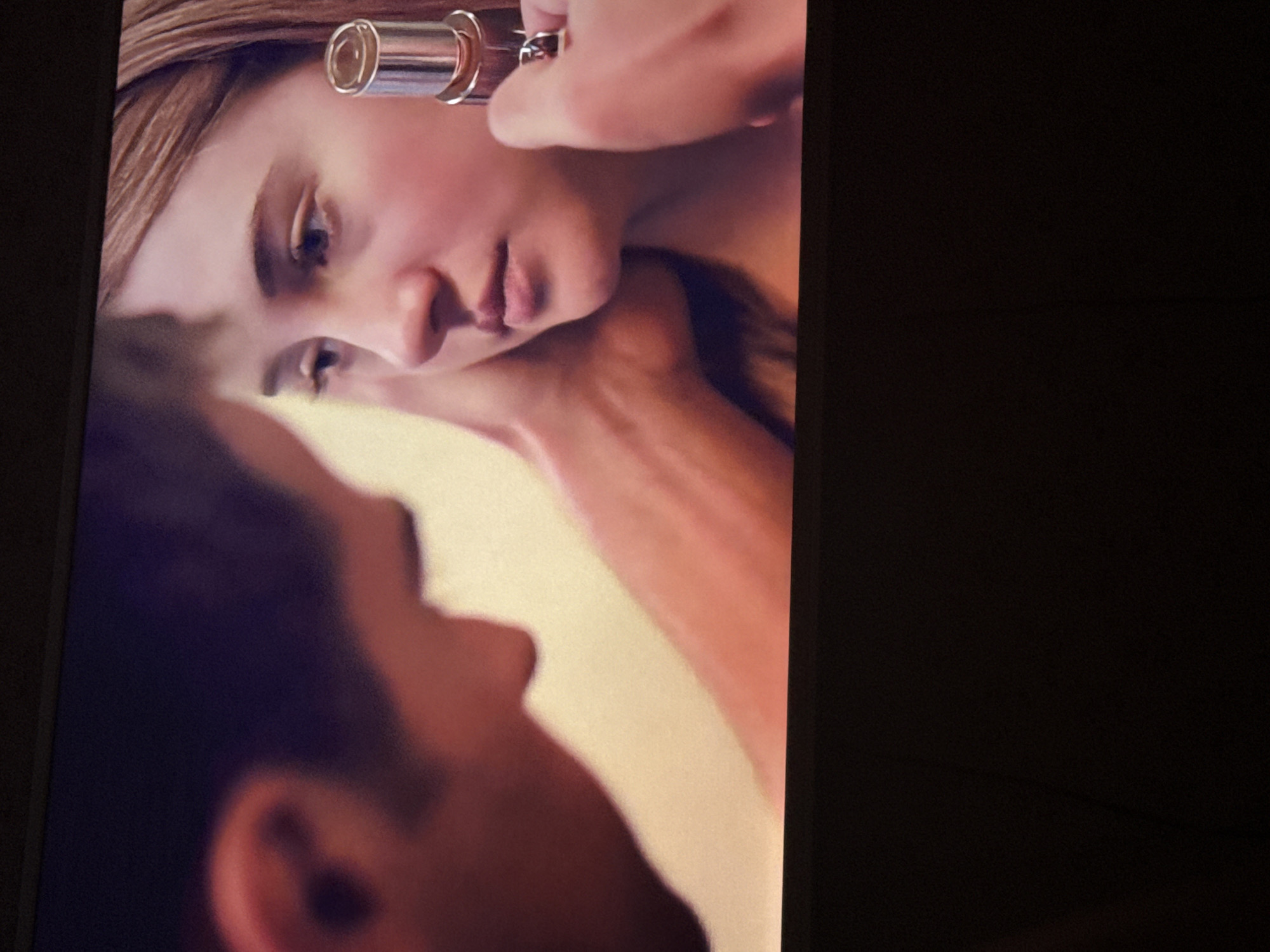 A close-up shot depicts a light-skinned woman in profile as she is being touched up with makeup. A tube of lipstick or concealer is positioned near her face, presumably for application. Part of a man's face is visible in the lower left corner of the frame. The background is a neutral color. There is insufficient information to determine the location is the city of Mytishchi, Russia.