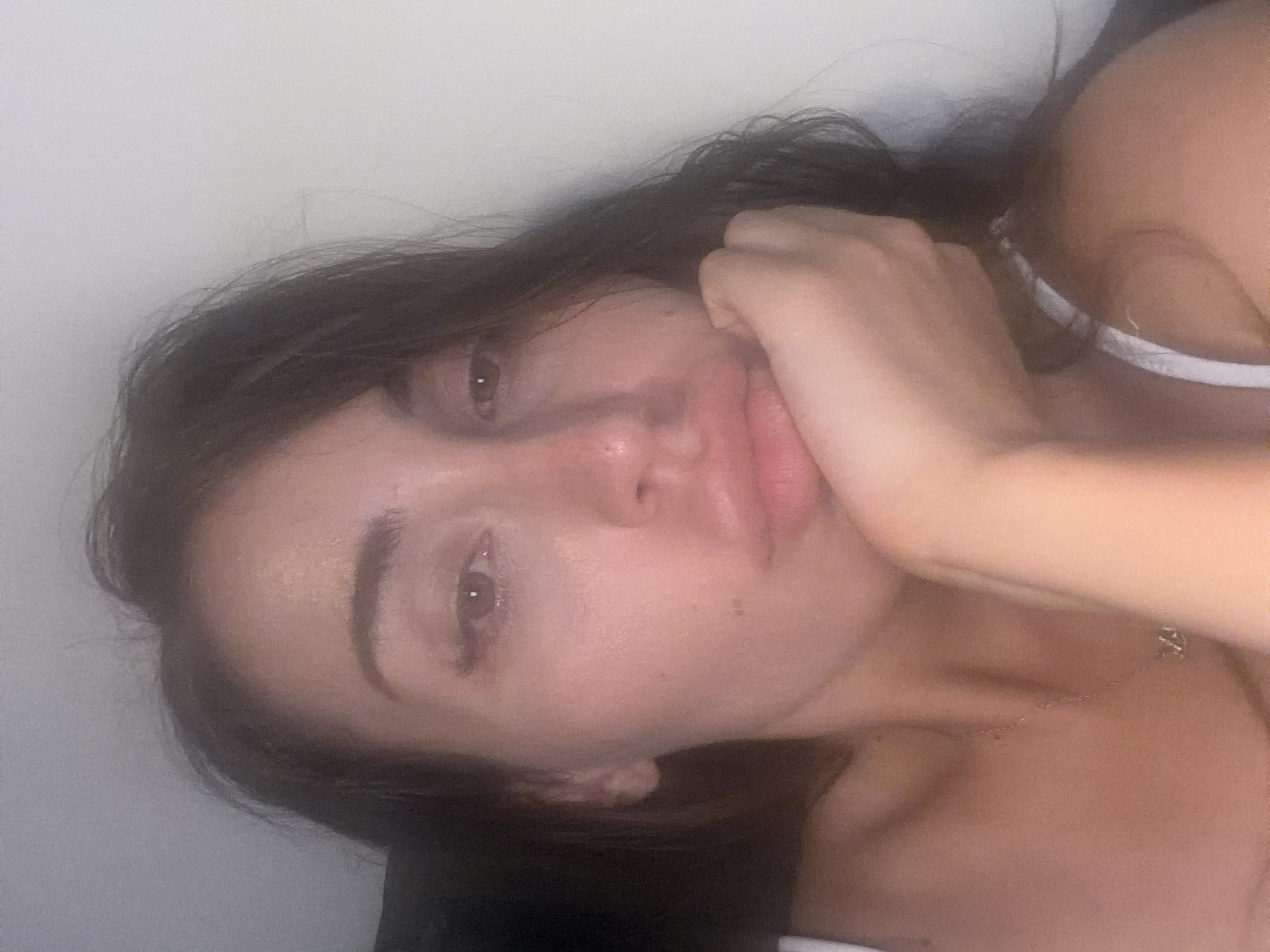 A close-up, dimly lit, over-the-shoulder selfie captures a young woman in Tersefanou, Cyprus. She is leaning her head on her hand, with her lips slightly parted. She has dark hair that frames her face, and her eyes are open, looking directly at the camera. She appears to be wearing a white top. The background is a plain, pale wall, and the lighting is soft and diffused. There is a delicate silver necklace visible around her neck.