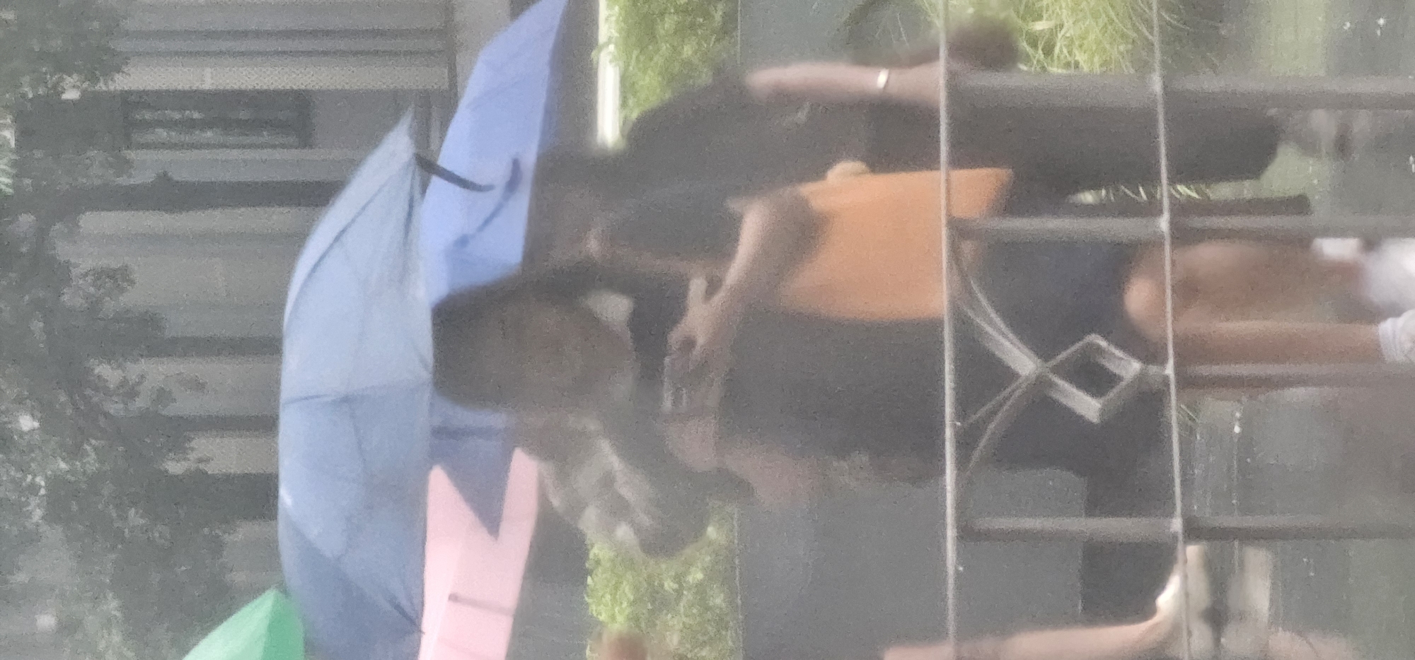 Two individuals are standing under umbrellas, one blue and one pink, in what appears to be a rainy day in Makati, Philippines. The person in the foreground, holding a phone, is wearing a dark top and an orange bag. The other individual, mostly obscured, is also holding an umbrella and wearing dark clothing. A metal fence is in the foreground, and lush green foliage is visible in the background, suggesting an outdoor urban setting. The lighting is diffused, consistent with overcast or rainy conditions.