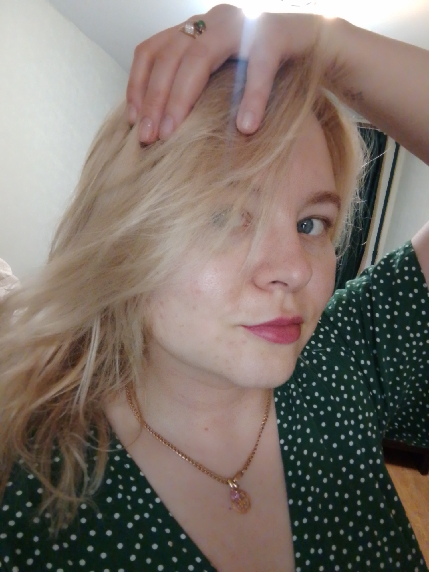 A woman is in a selfie-style shot, facing the camera with her left hand raised and touching her hair. She has blonde hair that is partially covering her left eye. She has light skin, pink lipstick, and blue eyes. She is wearing a green dress with white polka dots and a gold necklace with a pendant. Her right arm is visible, and there appears to be a small tattoo on her upper arm. She is wearing a ring on her left hand. The background is out of focus, showing a light-colored wall. There is no visible information to determine the location in Izhevsk, Russia.