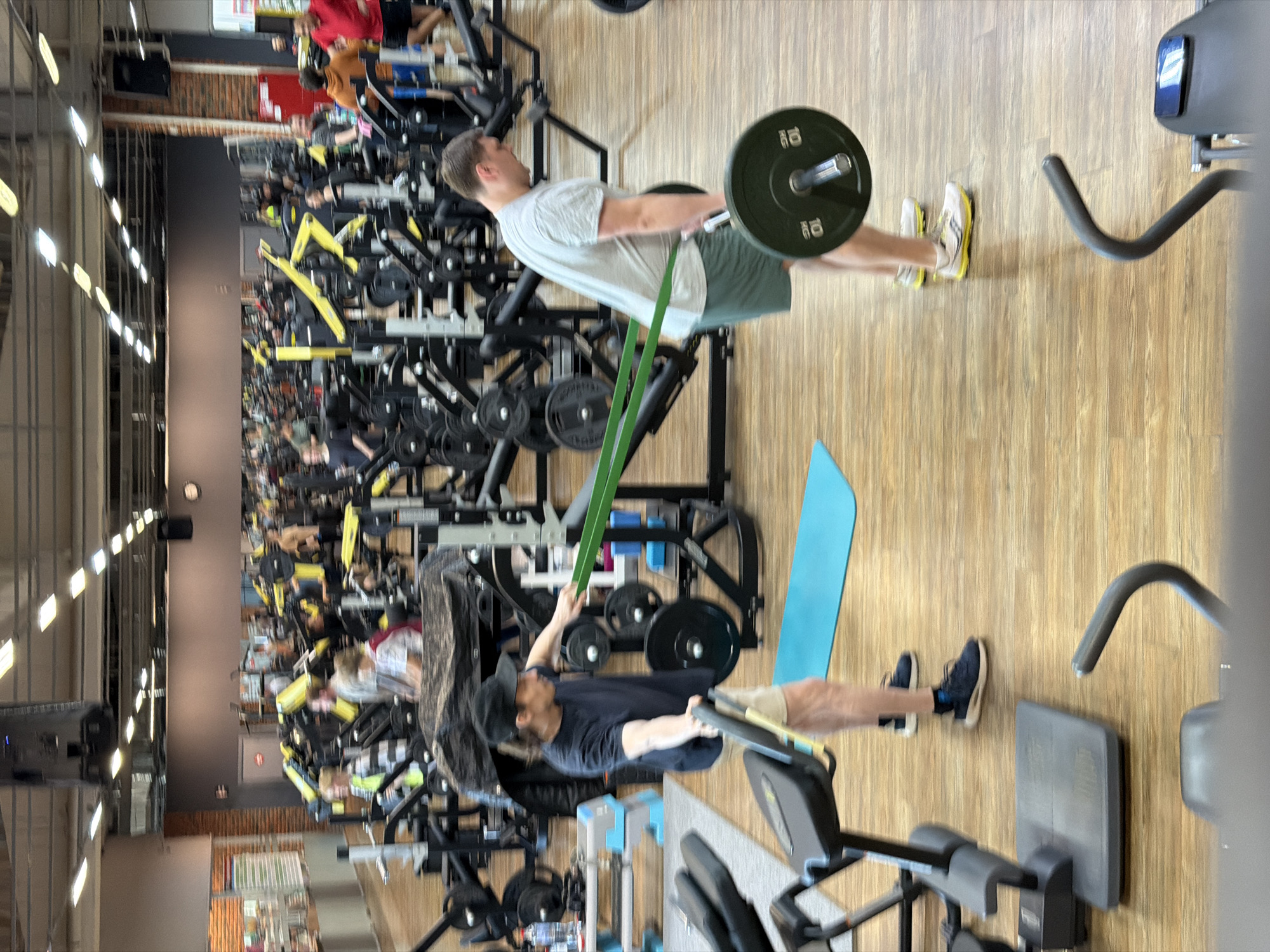 The image shows the interior of a gym, crowded with exercise equipment and people working out. Two men are prominently featured: one lifting weights using a resistance band, and the other performing a barbell exercise. The gym appears to be in Chaikovsky, Russia.