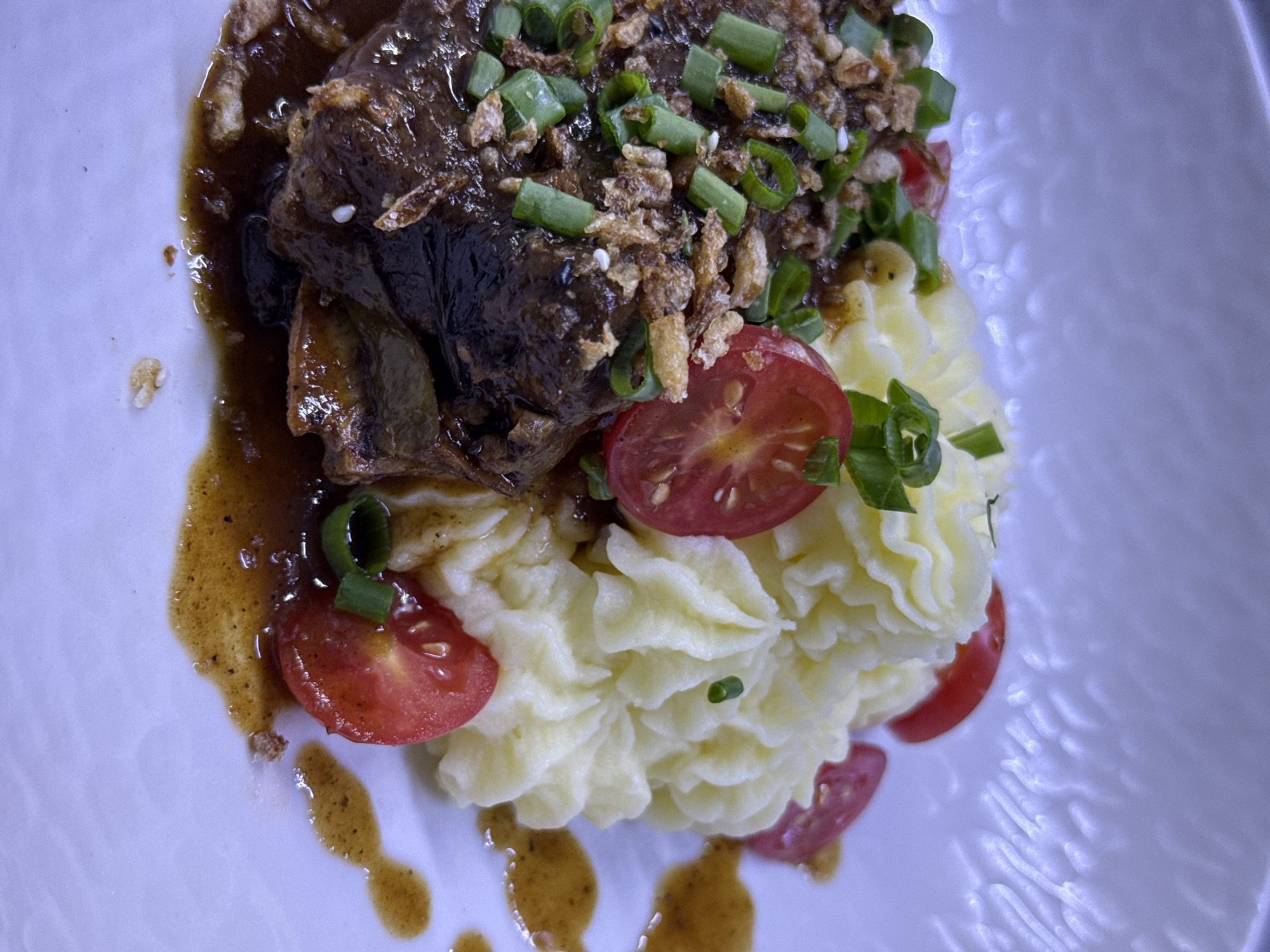 A close-up, overhead shot captures a meticulously plated dish on a white, textured plate. The dish features a dark, braised piece of meat, possibly short ribs, generously coated in a glossy, brown sauce. The meat is topped with chopped green onions and crispy fried shallots. To the right of the meat is a swirl of creamy mashed potatoes, artfully piped. Halved cherry tomatoes are scattered around the plate, some nestled into the potatoes and others alongside the meat. The sauce from the meat has dripped and pooled on the plate, adding to the rich presentation. The background is a soft, blurred white, suggesting a restaurant setting. The lighting is somewhat dramatic, with highlights on the glossy sauce and the creamy texture of the potatoes. There are no people or animals visible in the image. The location is not specified in the image.