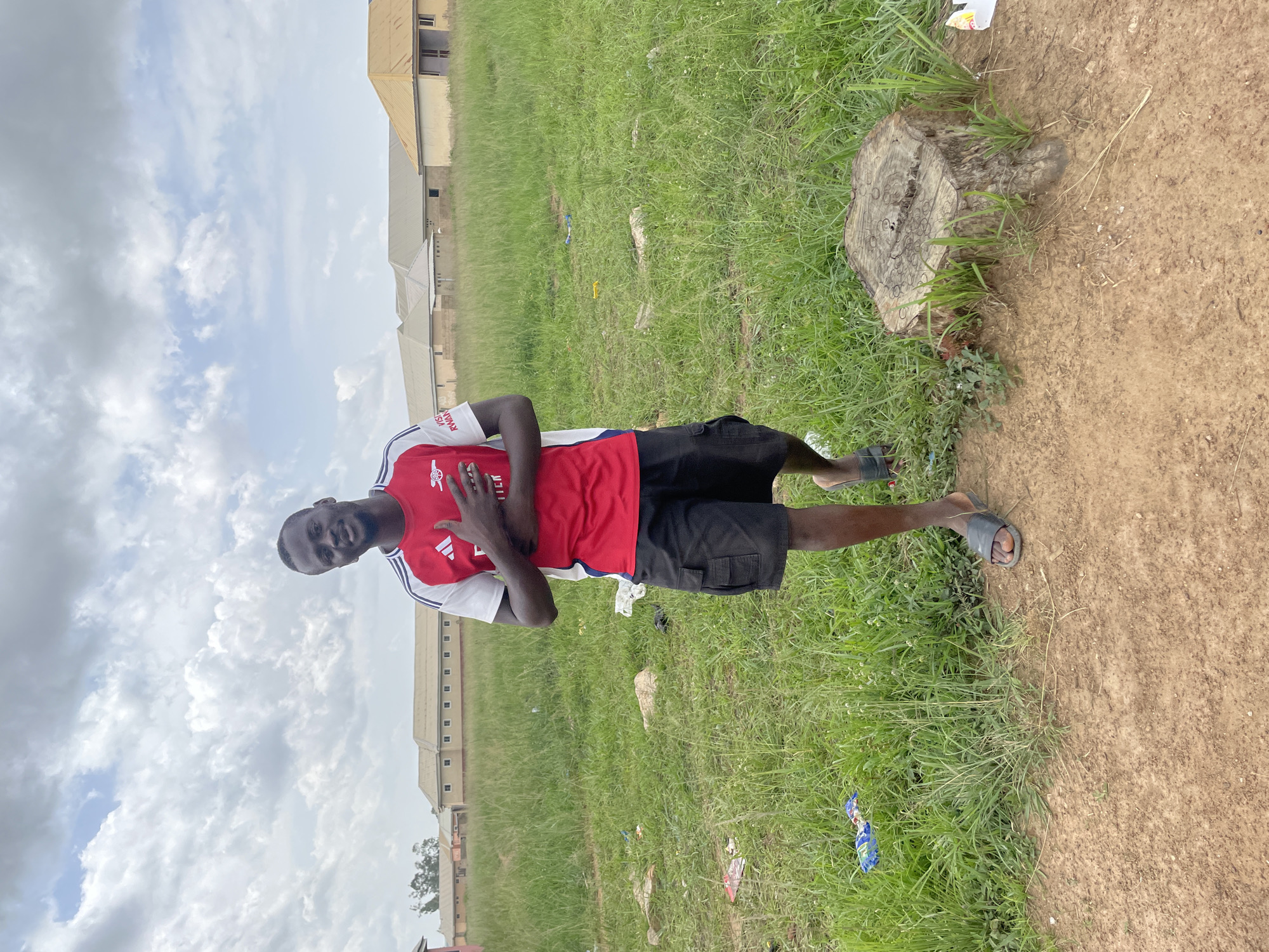 A young Black man, wearing a red and white short-sleeved jersey with black shorts and dark-colored sandals, stands in a grassy outdoor area. The man's hands are crossed over his chest, and he is looking towards the camera with a slight smile. His jersey features a white collar and white stripes down the sleeves, with a crest on the left side of his chest. The background consists of a grassy field, a dirt path with a tree stump, and several buildings in the distance. The sky is filled with white clouds. The image appears to be taken during the daytime. There is some litter visible in the grass. A logo resembling a shield with an animal is visible on the left side of the man's shirt. Text on the shirt is partially visible and reads "VISIT RW..."