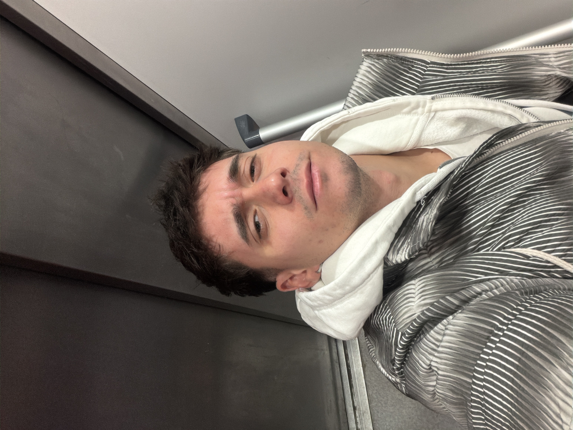 A man with dark hair and stubble is posing for a selfie in what appears to be an elevator. He is wearing a white hoodie underneath a black and white striped jacket. The elevator interior consists of gray panels and a metal handrail. The location cannot be determined from the image alone; the city of Sofia, Bulgaria, cannot be verified.