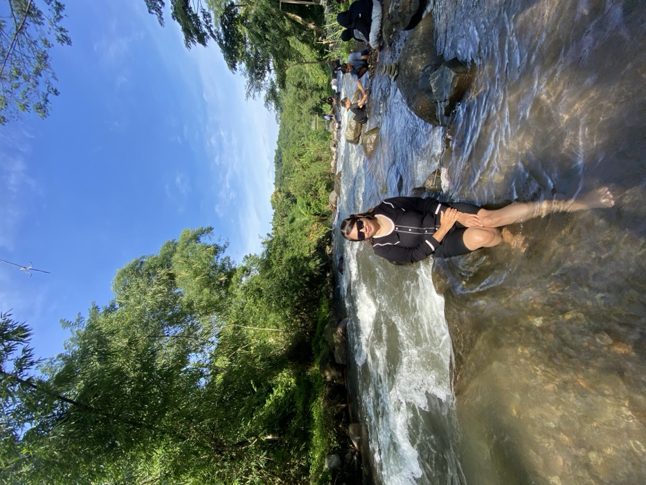 A woman in a black dress and sunglasses sits on a rock in a river, with her legs submerged in the water. Other people are seen in the background, sitting on rocks near the riverbank. Lush green trees and vegetation surround the river, and a clear blue sky is visible above. The water is flowing rapidly, creating white water near the woman.