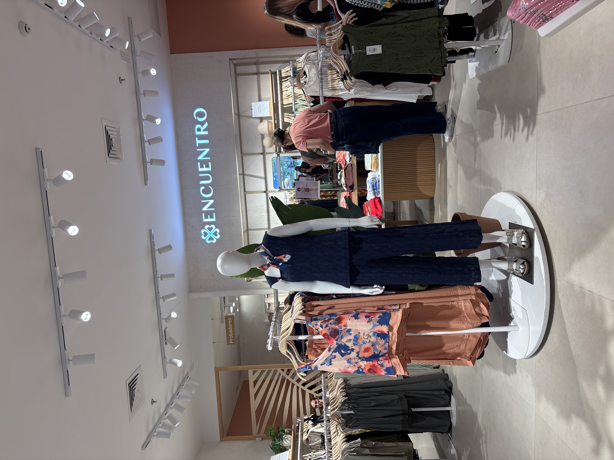 The image shows the interior of a clothing store. In the foreground, a mannequin is displayed wearing a dark blue sleeveless top with a patterned scarf and matching dark blue patterned pants, along with silver sandals. To the left of the mannequin, several garments are displayed on racks, including a sleeveless top with a floral print. In the background, the store's name, "ENCUENTRO," is visible in illuminated lettering on the wall. Several shoppers are browsing the clothing racks and displays. The store appears to be well-lit with track lighting on the ceiling. The flooring is tiled. The overall atmosphere is that of a modern retail environment. There is no indication in the image of the city of Orihuela, Spain.
