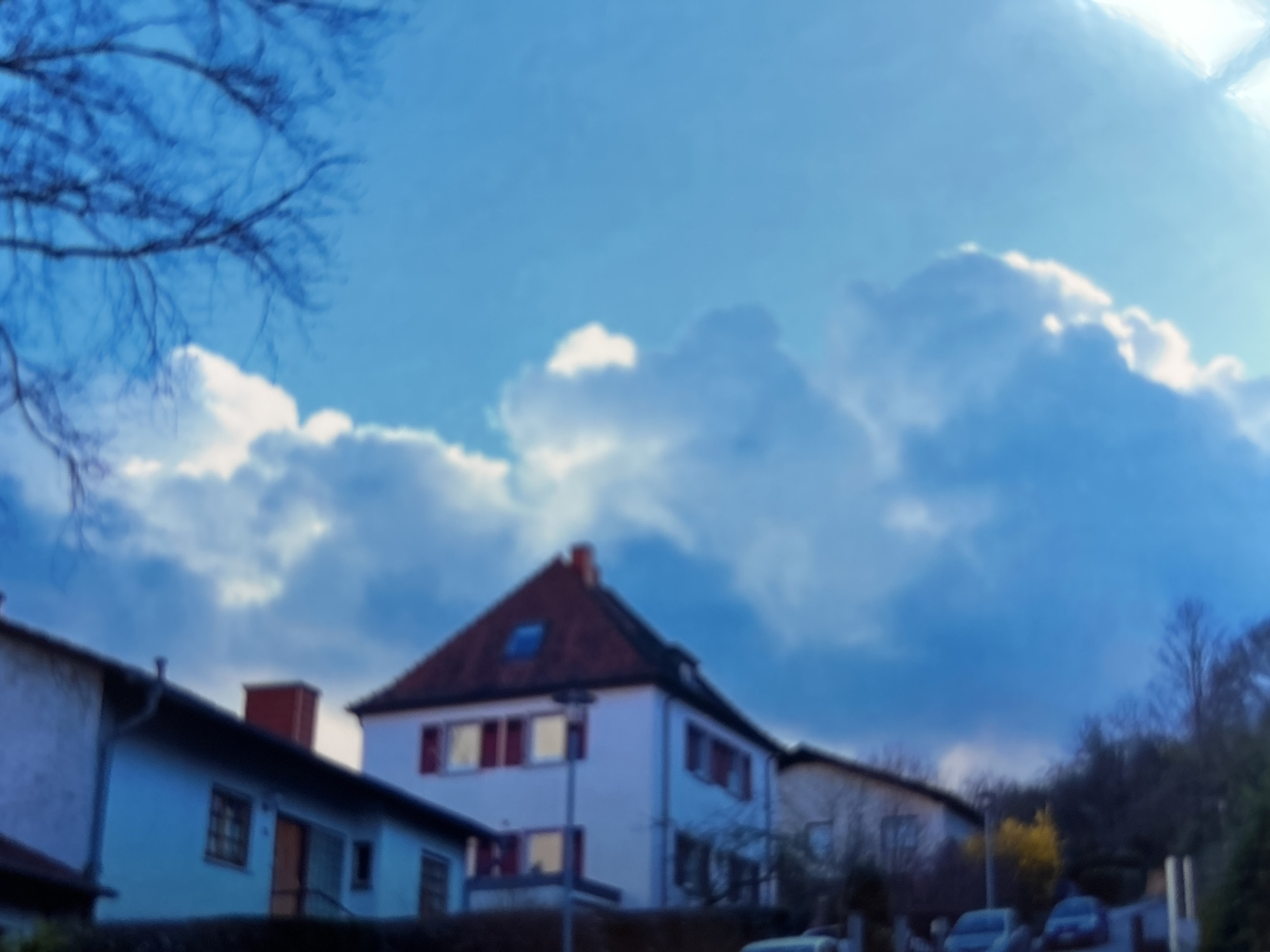 The image depicts a residential building against a bright blue sky with scattered clouds. The building appears to be a multi-story structure with a red-tiled roof and white walls. Several windows with red frames are visible on the facade. Bare tree branches are seen in the upper right portion of the frame, suggesting it might be a colder season. The scene is shot from a low angle, looking upwards towards the sky and the top of the building. The image is slightly out of focus, giving it a soft, almost dreamlike quality. The location is described as Unknown, Unknown.