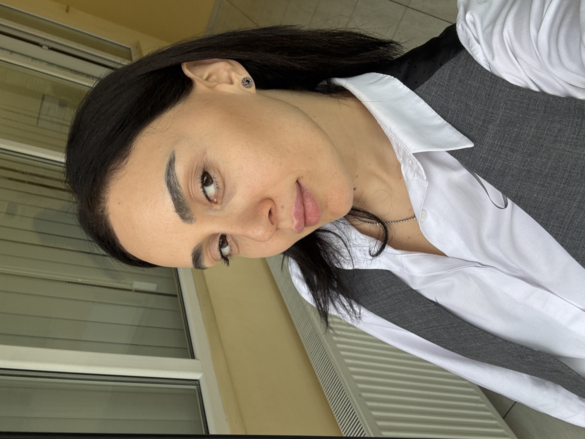 A medium shot features a woman with dark hair, styled straight, and defined eyebrows. She is wearing a white button-down shirt with a dark grey vest. A thin silver necklace is visible around her neck. Her skin appears smooth, and she is wearing stud earrings.
The backdrop includes a light yellow painted wall and a white-framed window with closed blinds. The location is suggested to be in the city of Tbilisi, Georgia, based on the prompt.