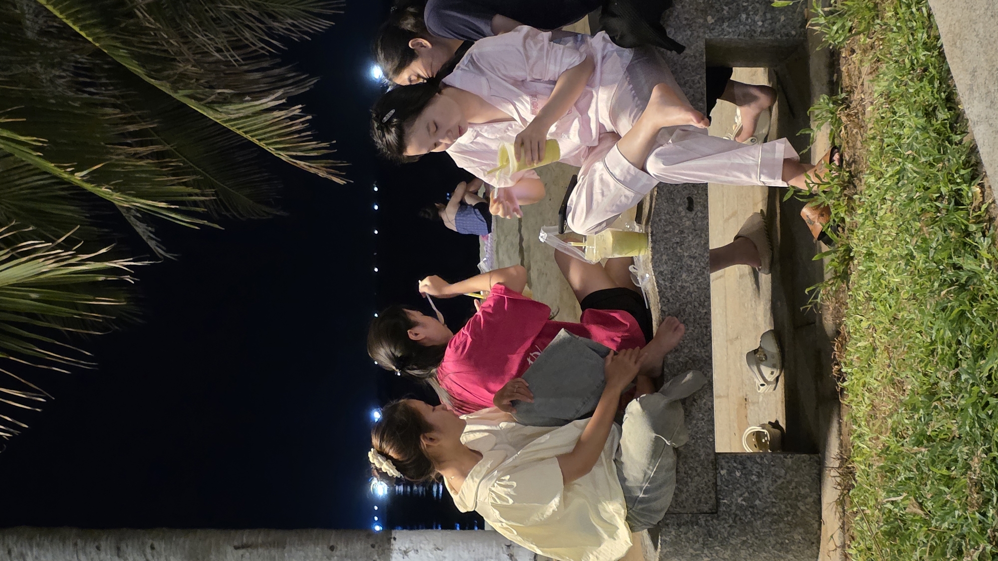 The image depicts a group of four young women in Nha Trang Ward, Vietnam, gathered in an outdoor setting at night, close to what appears to be a beach based on the palm tree and water visible in the background. The women are positioned along the edge of a concrete structure with lush green grass growing adjacent to it.
One woman is seated wearing pink pajamas and pouring a beverage from a plastic cup into another similar cup. To her left is another woman wearing a dark top. Below the woman in pink pajamas is another woman sitting in a pink shirt and shorts, appearing to eat or stir something in her cup with a spoon. She's facing another woman in a white shirt and gray pants. These two seem to be interacting with each other.
The scene is illuminated by overhead lighting and possibly some lights along the shoreline. The environment appears casual and informal.