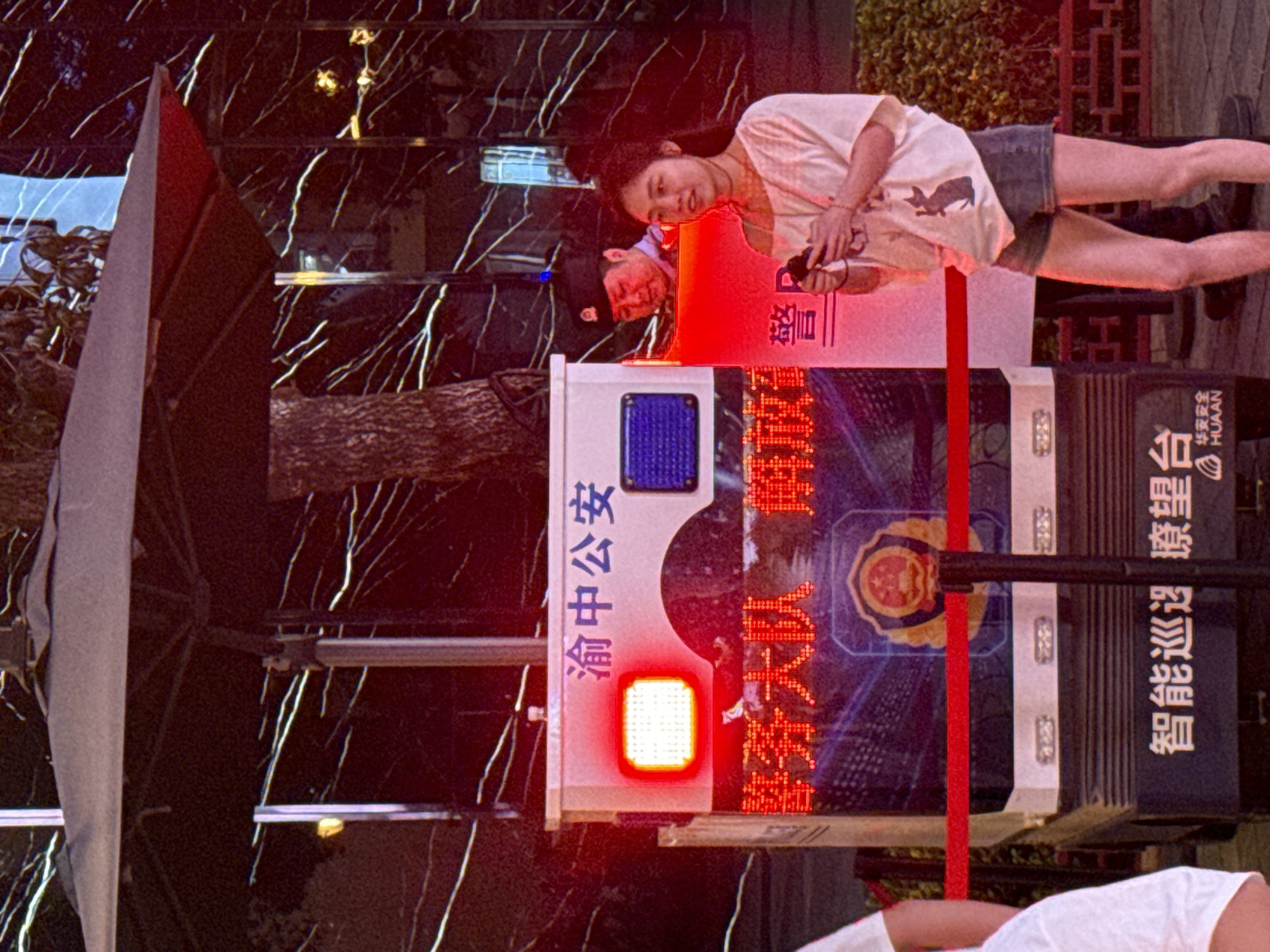 A street scene in Yuzhong District, China, features a police kiosk with glowing red and blue lights and Chinese characters. A young woman in a white t-shirt and denim shorts stands to the right of the kiosk, holding a smartphone. Behind her, a uniformed police officer is visible within the kiosk. Red tape cordons off the area in front of the kiosk. The background is a dark, textured wall with white lines, possibly a building facade.